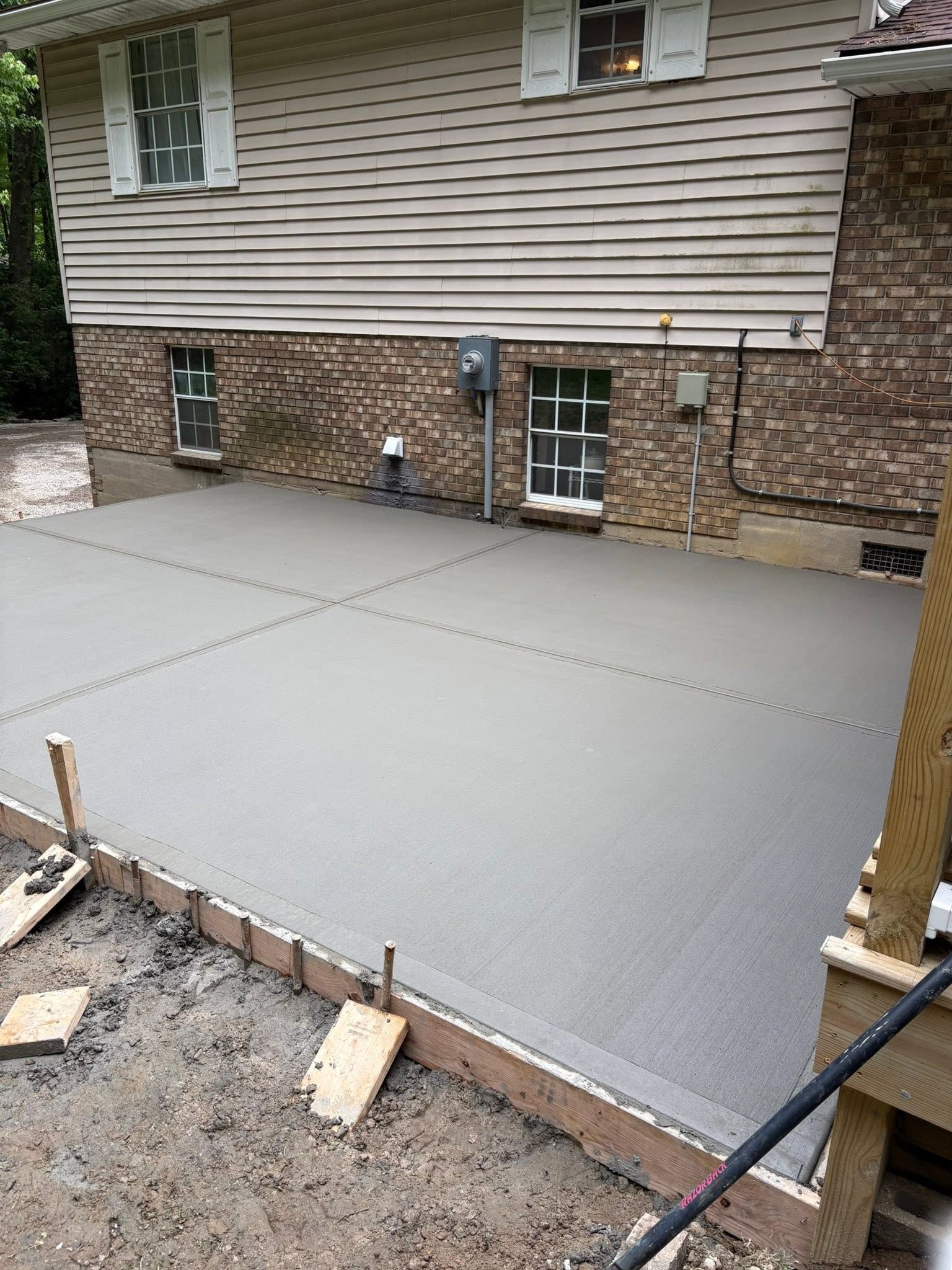 Freshly poured concrete patio slab beside a house with stone foundation and siding, forms visible at the edges