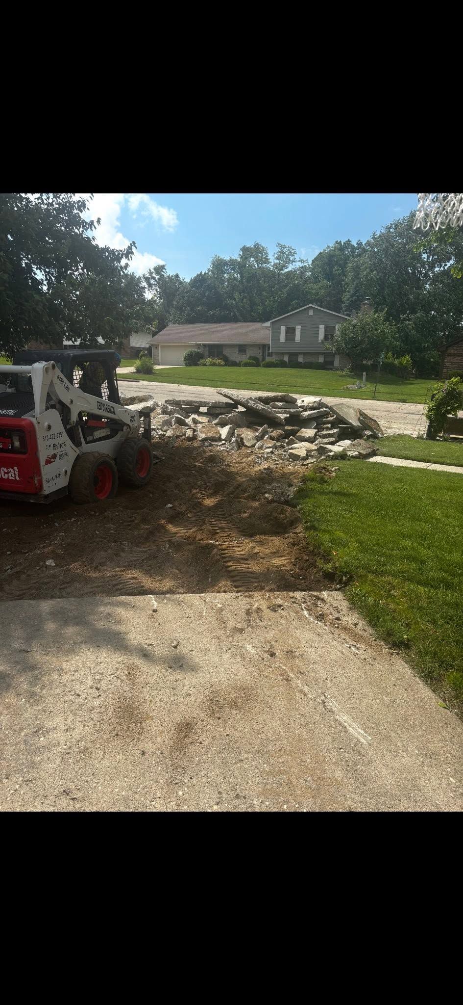 Construction equipment beside a gravel path near a building, with a large pile of rocks and trees in the background