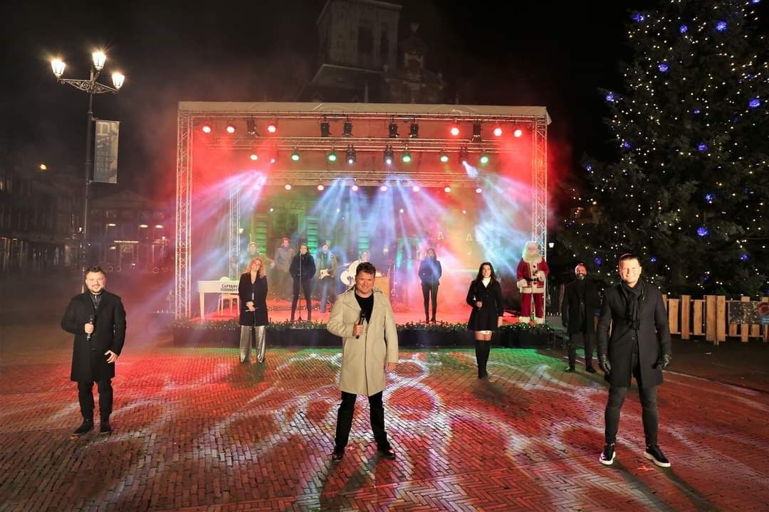 Vijf artiesten op een verlicht buitenpodium 's nachts met rode en blauwe lichten en vallende sneeuw.