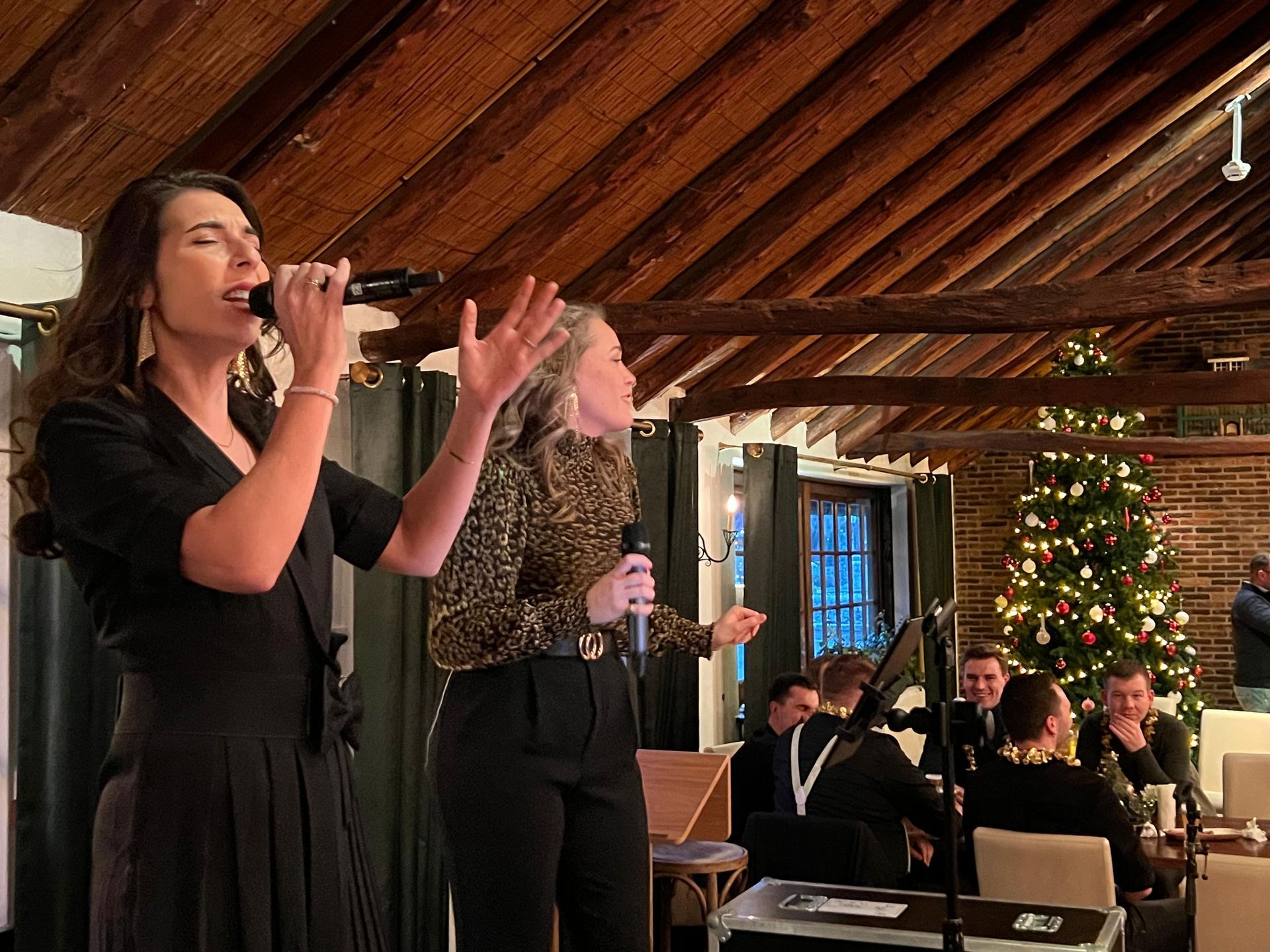 Twee vrouwen zingen op een podium in een rustieke zaal, met een verlichte kerstboom en zittende gasten in de buurt.