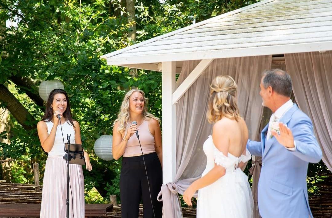 Bruid en man in blauw pak staan ​​tegenover twee vrouwen die foto's maken naast een wit prieel buiten.