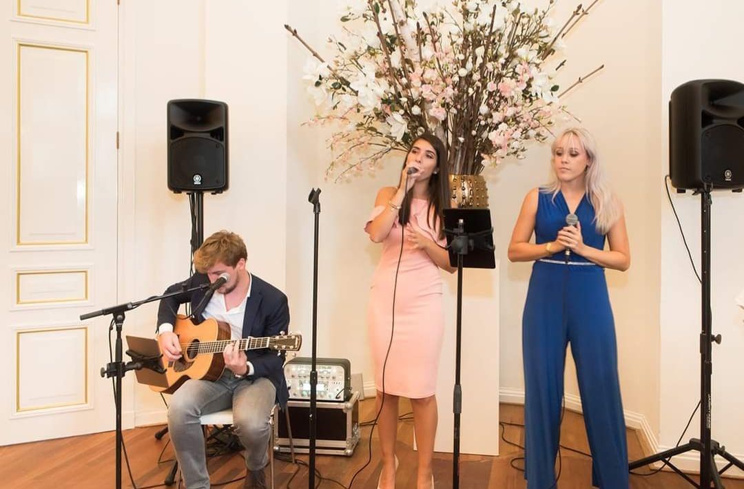 Drie muzikanten treden op in een lichte zaal; één speelt gitaar, twee zingen via microfoons.