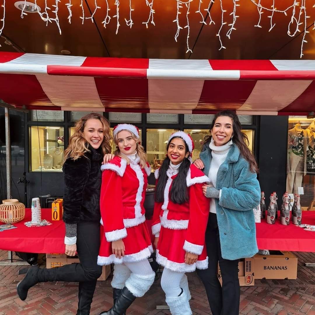 Vier vrouwen in kerstmanpakken poseren bij een feestelijke marktkraam met een rood-wit afdak en kerstverlichting.