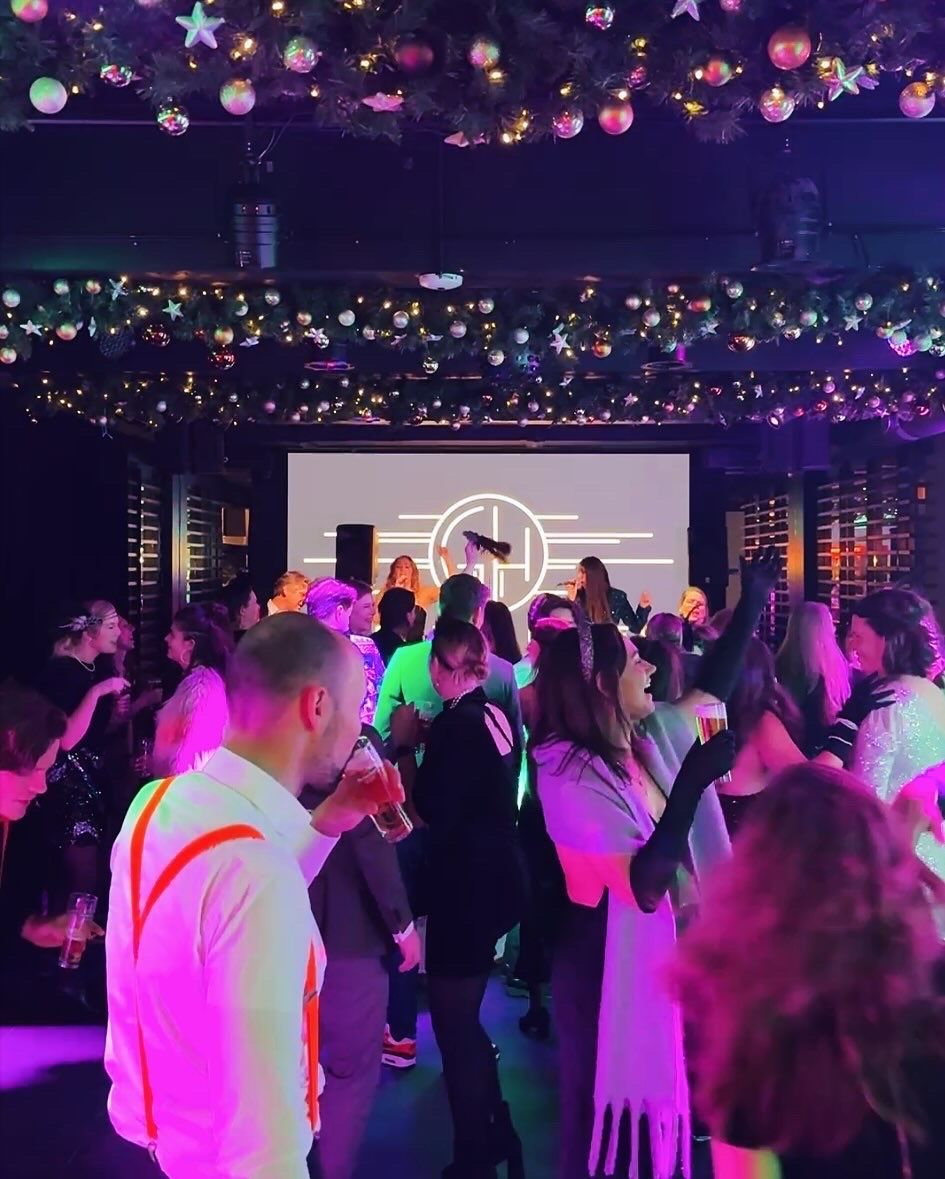 Een druk feest in een donkere zaal met paarse verlichting en een verlicht podium op de achtergrond.