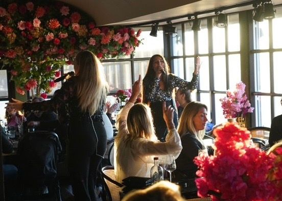 Een evenement binnenshuis vol bloemen, met vrouwen die zitten en staan ​​bij ramen waar het zonlicht doorheen schijnt.