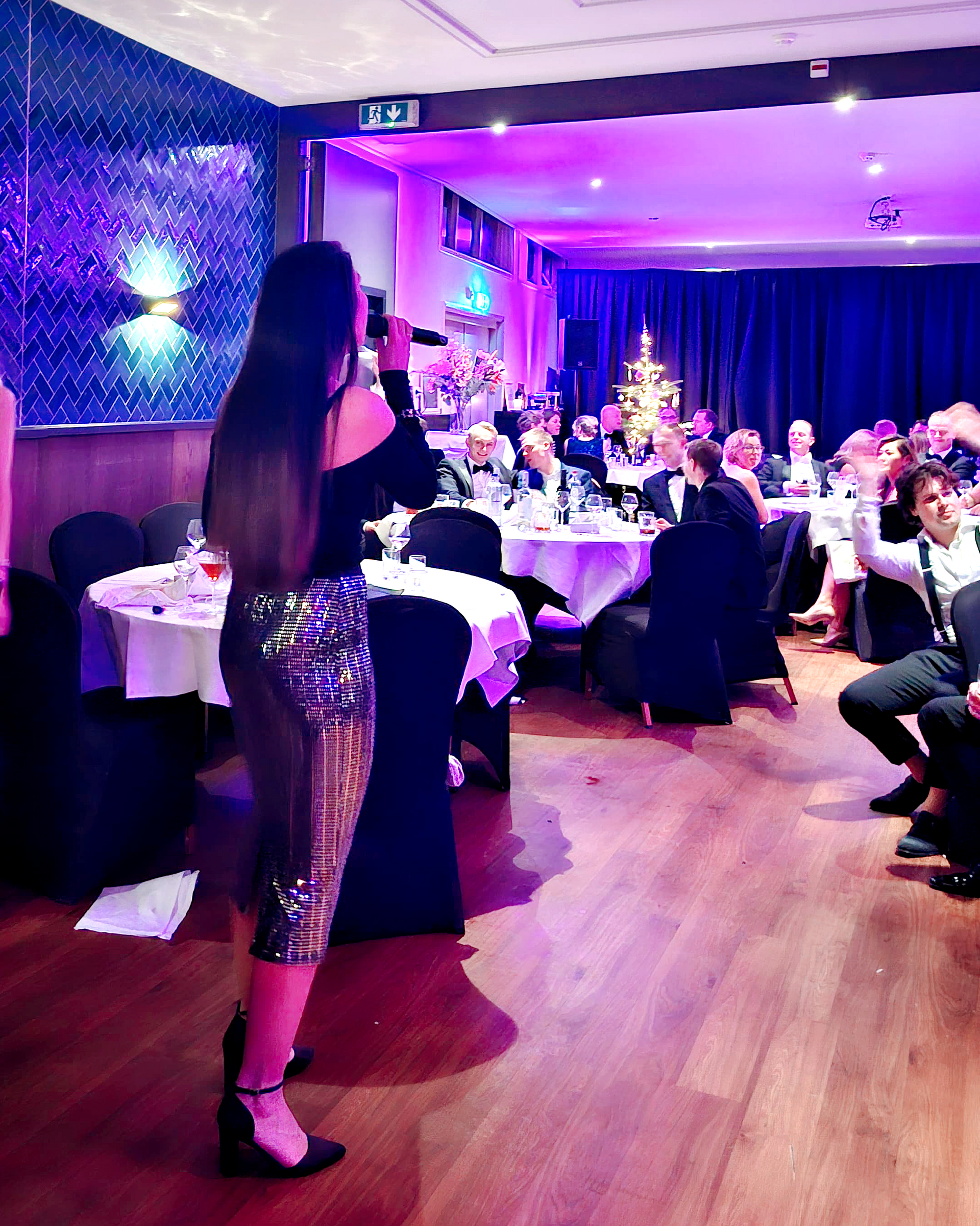 Een gast in een glinsterende legging spreekt tot de zittende gasten tijdens een evenement in een paars verlichte feestzaal.
