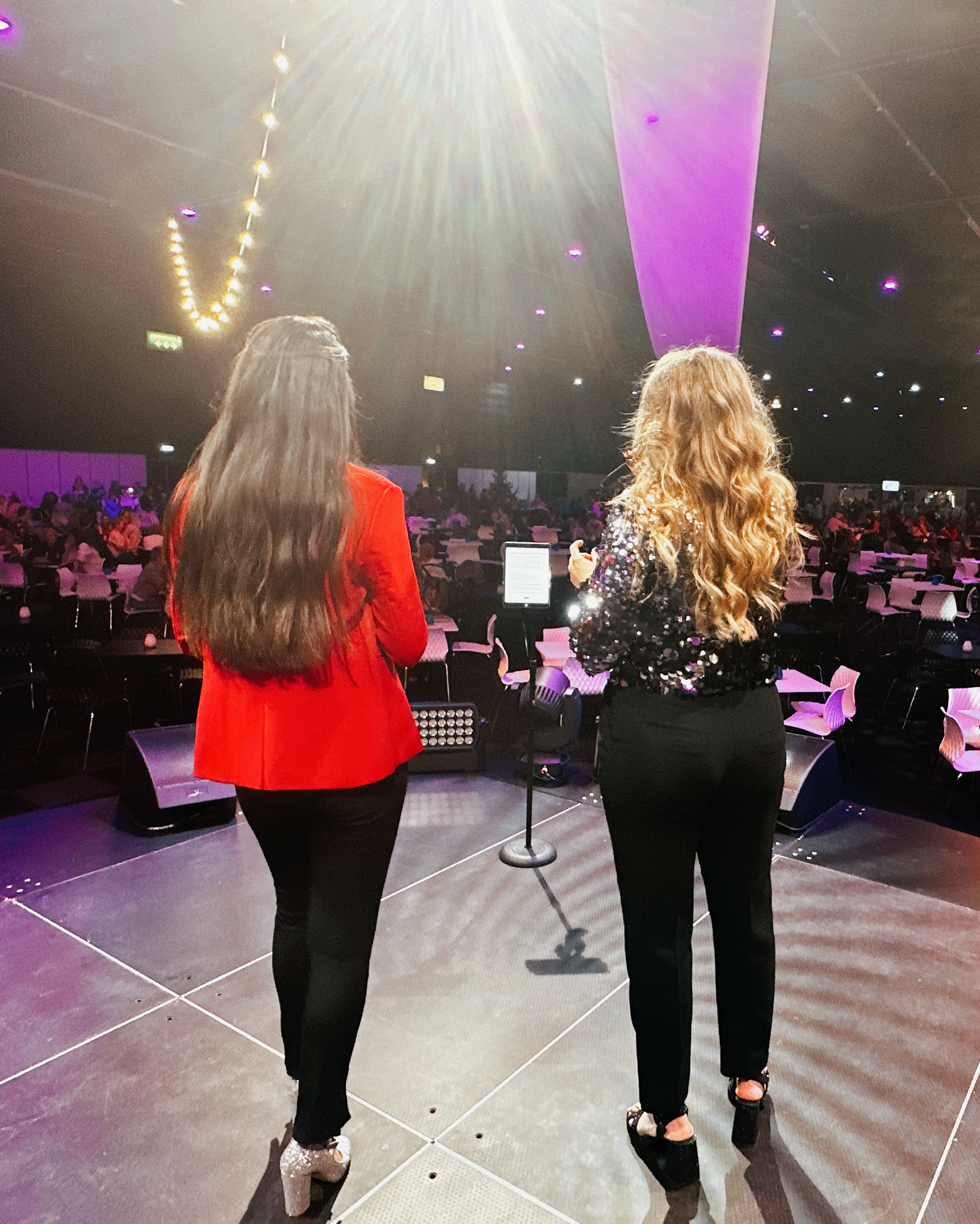 Twee vrouwen staan ​​op het podium tegenover een groot publiek, de een in het rood en de ander in het zwart, onder fel concertlicht.