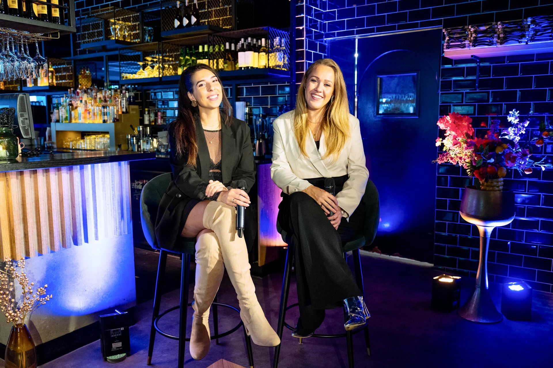 Zangduo Remedy zit op barkrukken in een lounge of nachtclub met neonverlichting, met drankjes en kleurrijke verlichting.