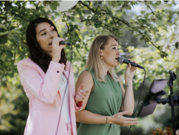 Zangduo Remedy treedt buiten op met microfoons onder lommerrijke bomen, de een in roze en de ander in groen.