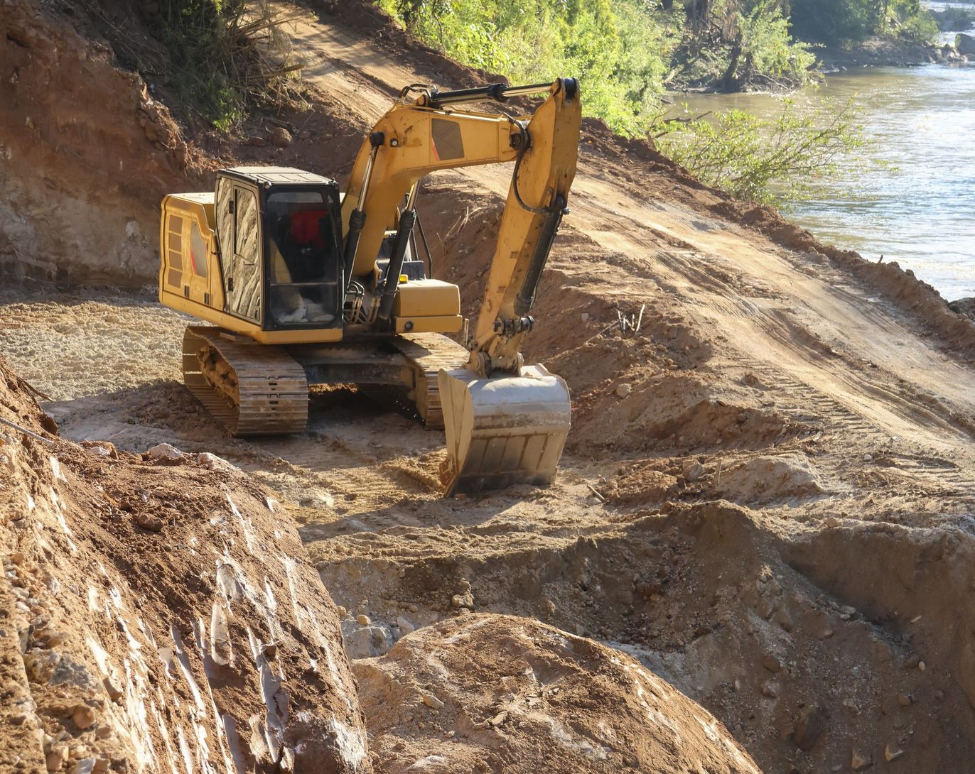 Yellow excavator on a dirt slope beside water, digging earth near the shoreline.