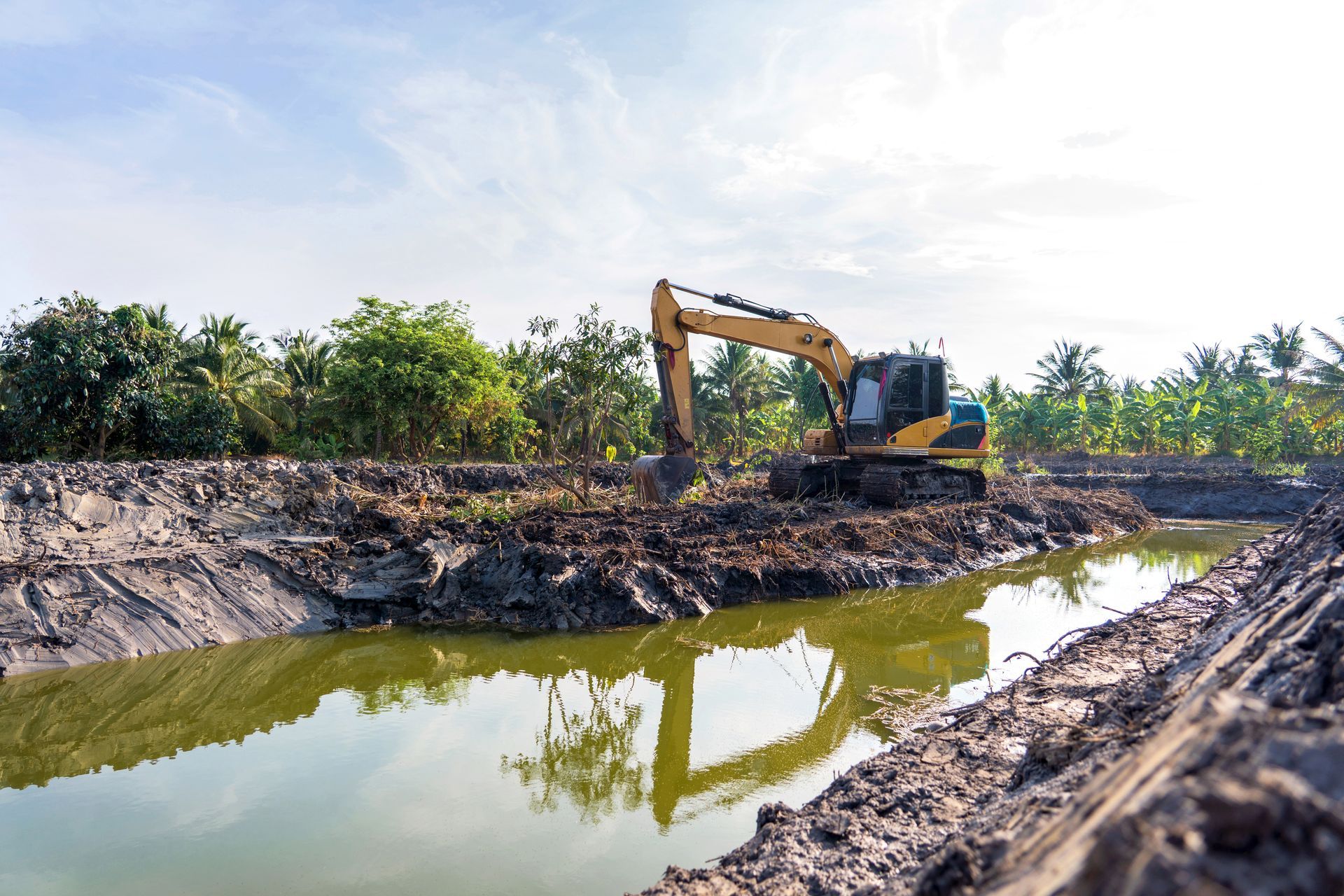 Excavator digging a muddy canal beside a narrow waterway under a bright sky