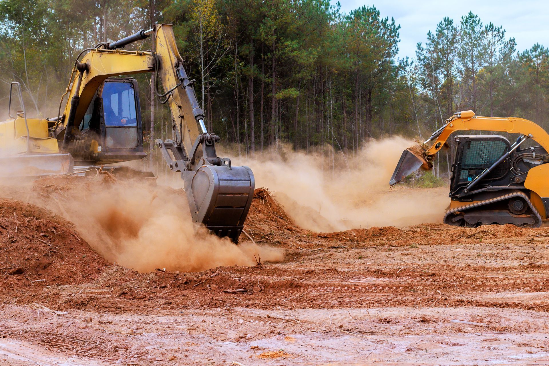 Two excavators digging in red dirt, kicking up dust in a wooded construction site.