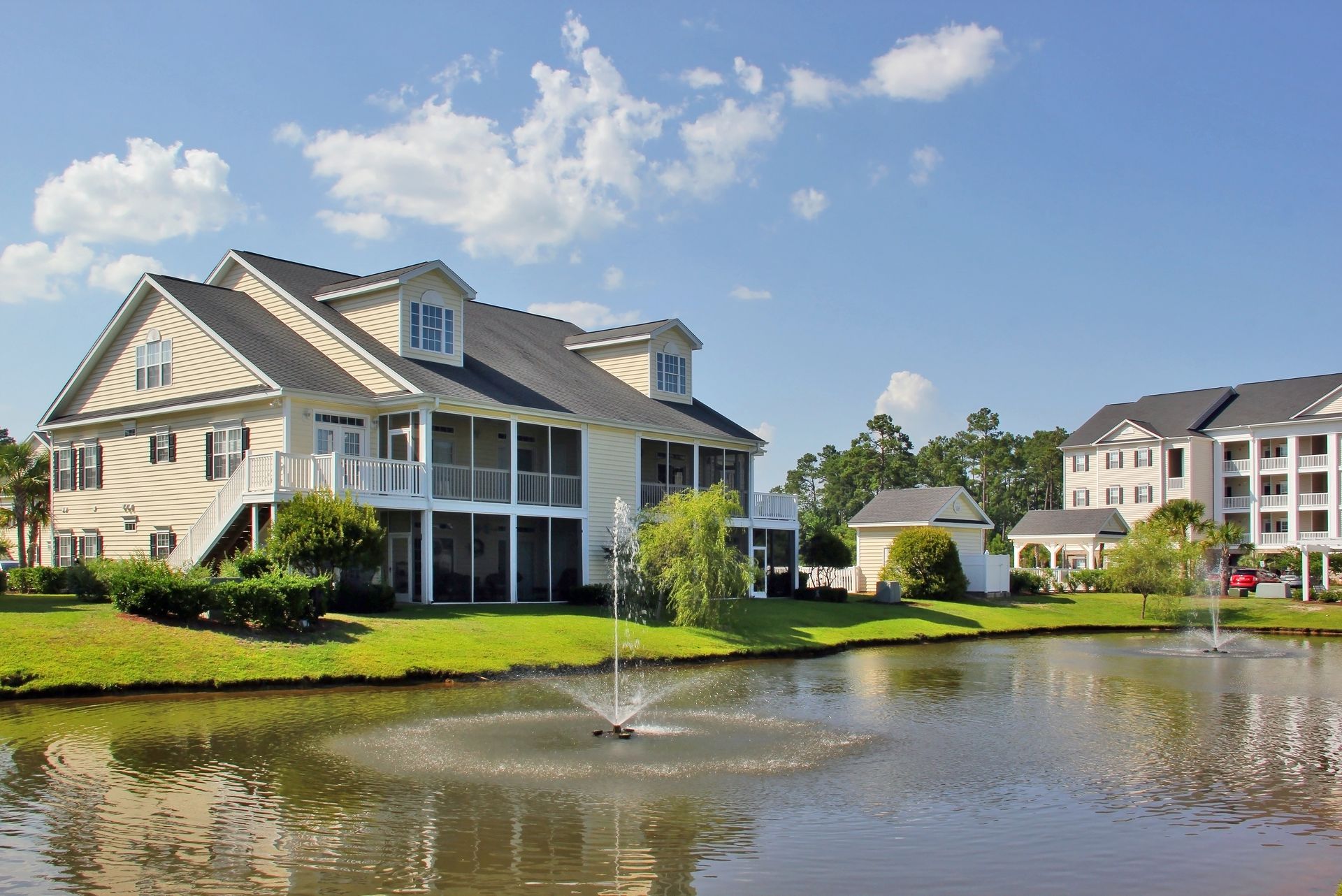 Large lakeside house with a fountain, blue sky, and neighboring homes in a suburban neighborhood