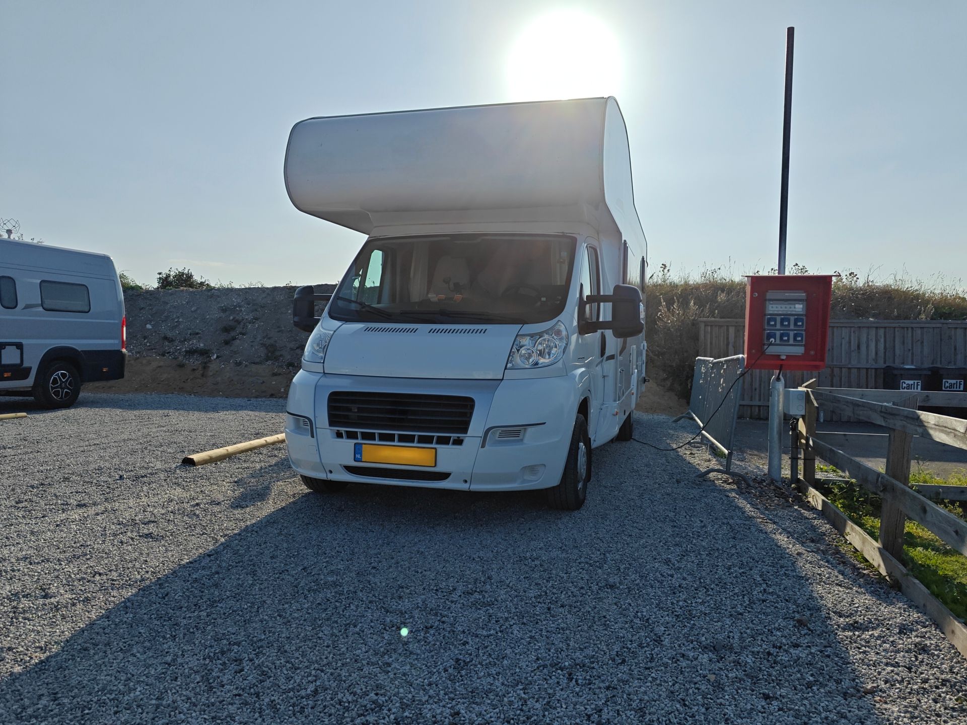 White camper van parked on gravel by a gate, with another vehicle and bright sun in the background