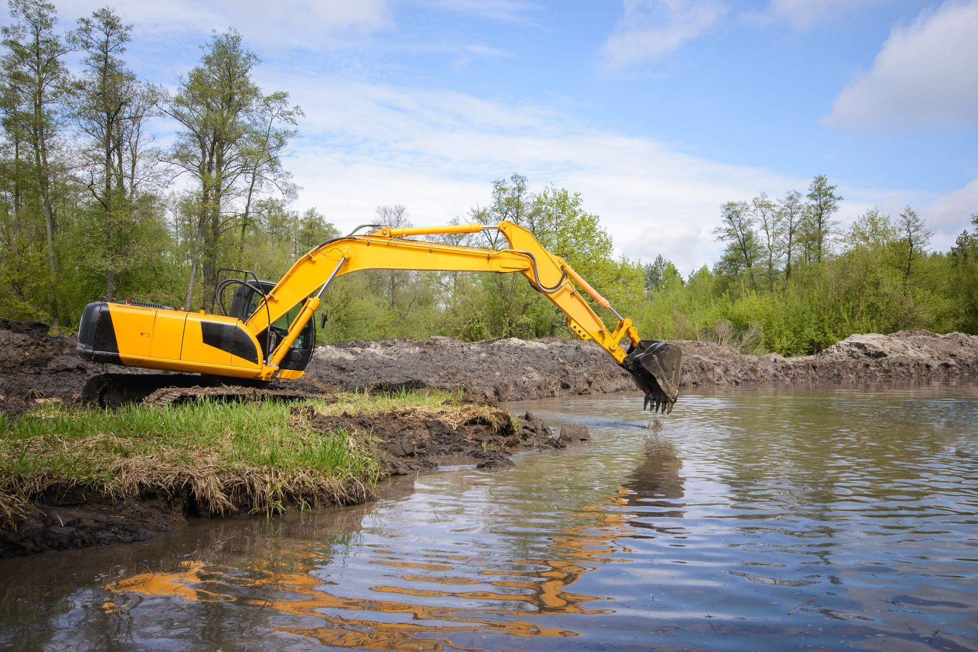Yellow excavator reaching into a riverbank beside a wooded shoreline under a blue sky