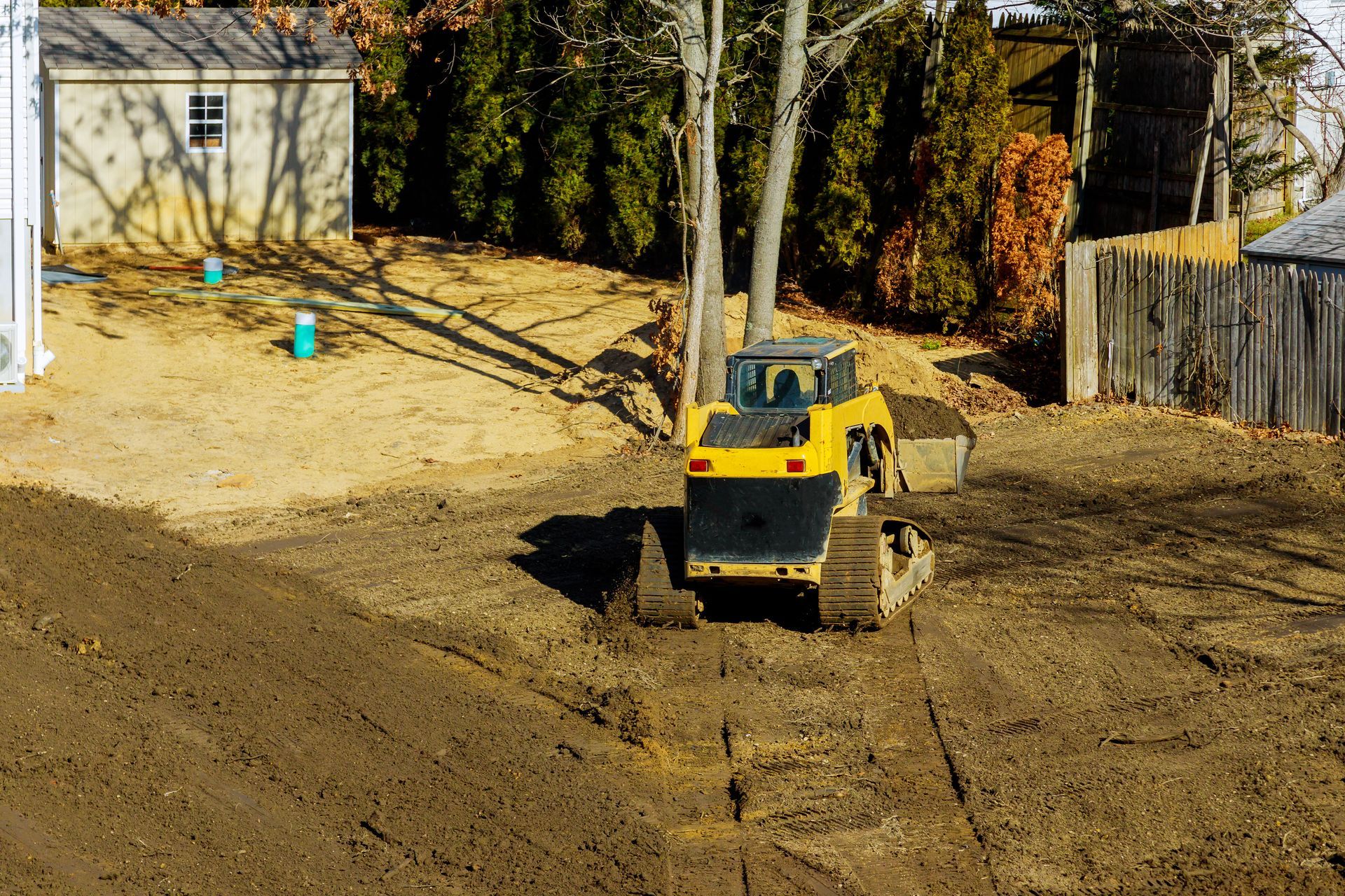 Yellow skid steer on a dirt lot near a garage and trees, with a wooden fence in the background