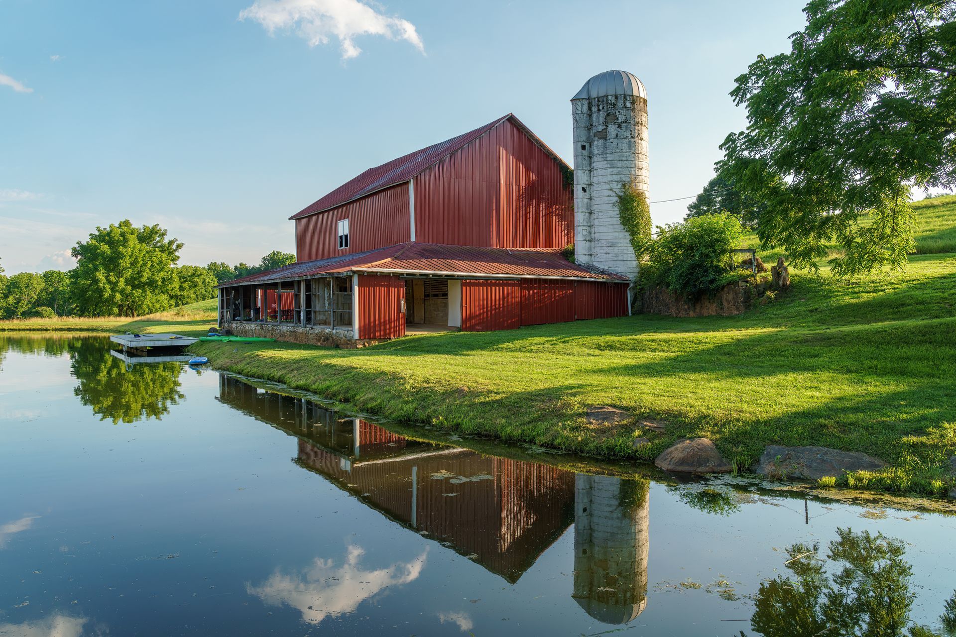 Red barn and silo beside a calm pond, with trees and their reflections in the water