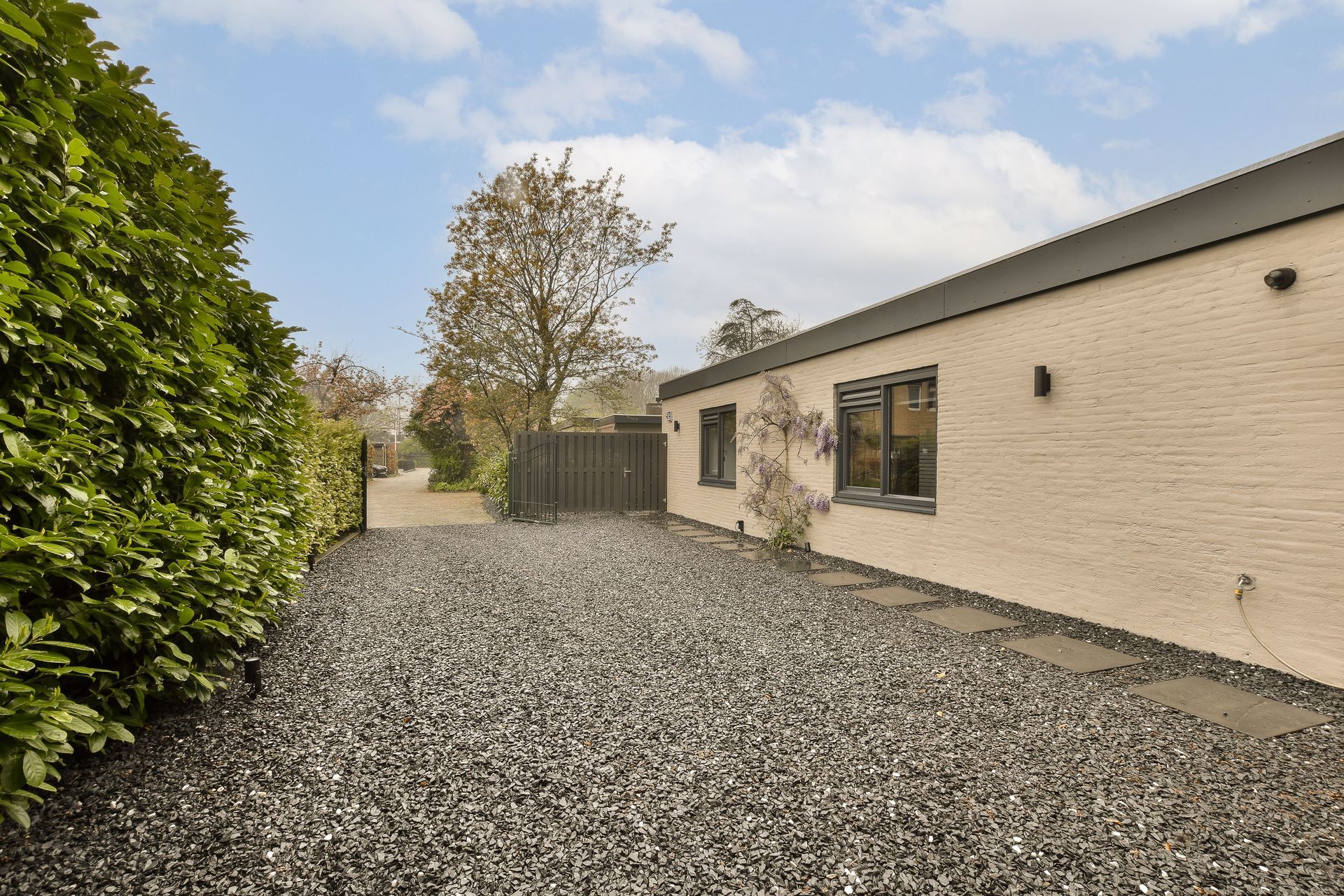 Gravel courtyard beside a white brick building with windows and a hedge under a cloudy sky