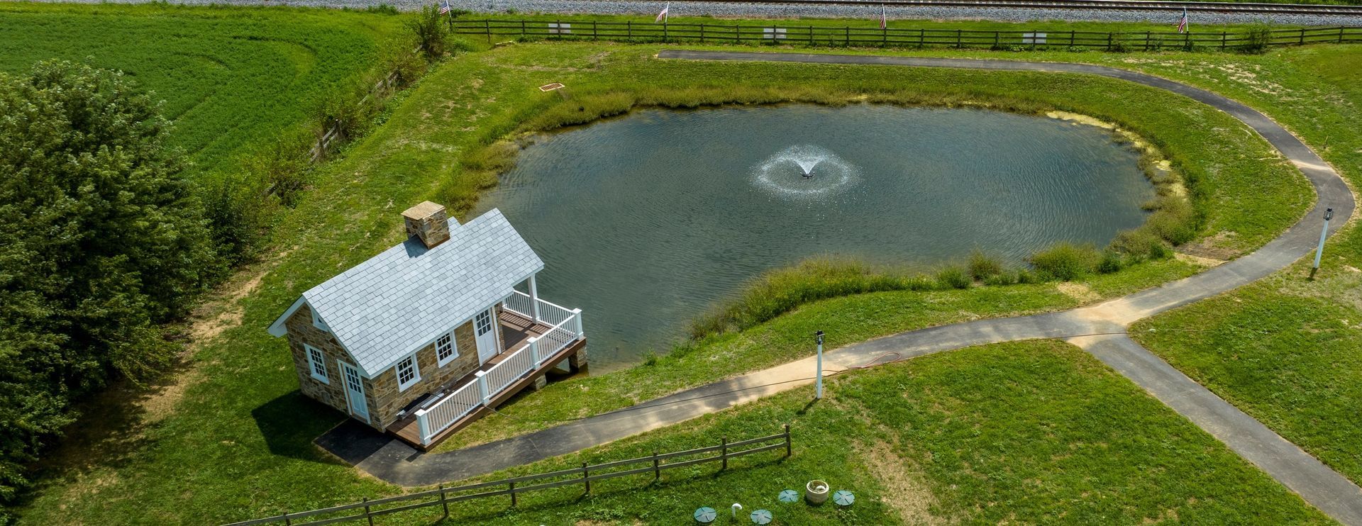 Aerial view of a small house beside a heart-shaped pond with a curving dirt road and green fields