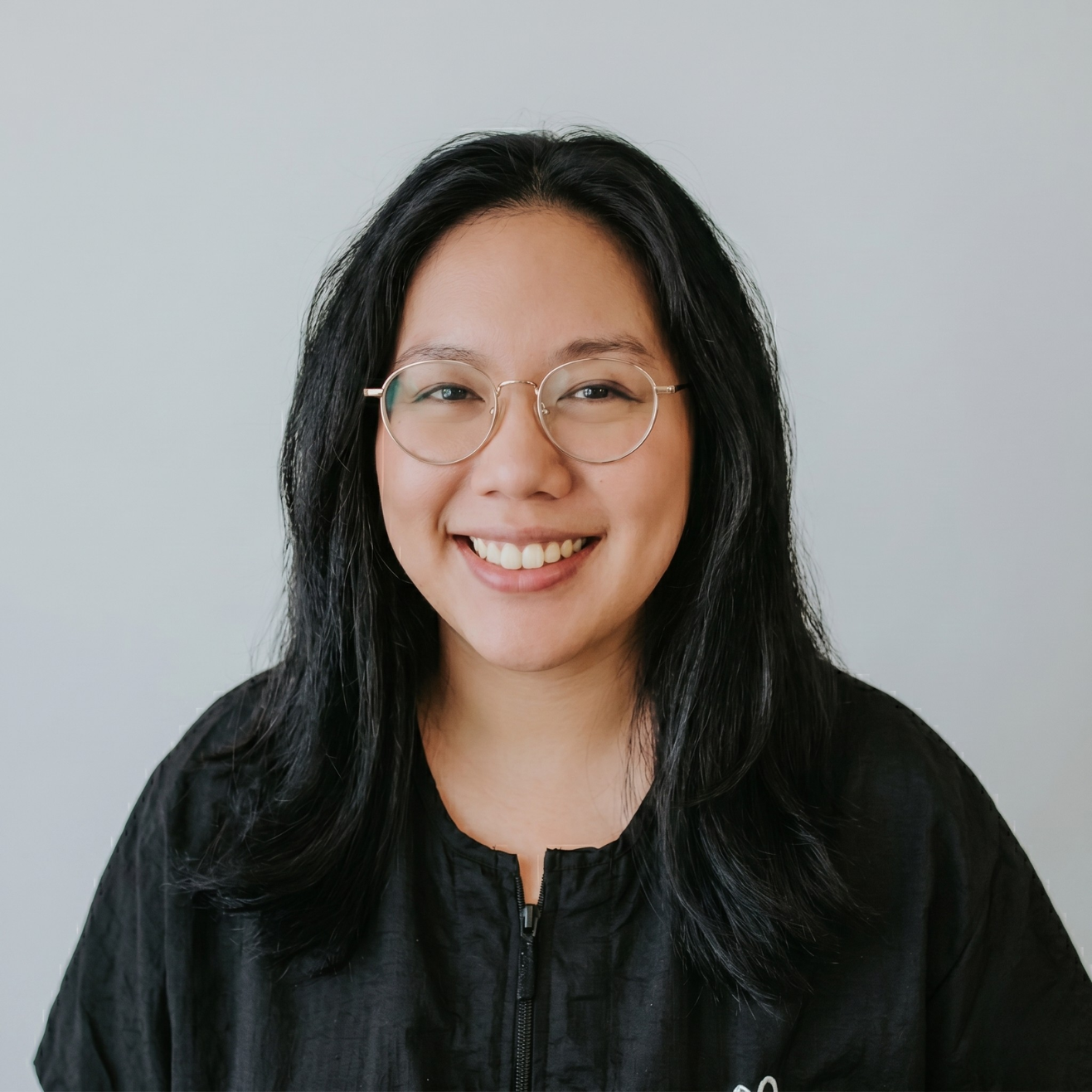 Smiling woman with glasses and long black hair, wearing a black top against a light gray background