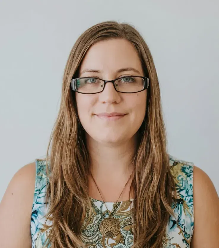 Portrait of a smiling person with glasses and long hair, wearing a patterned sleeveless top on a light background