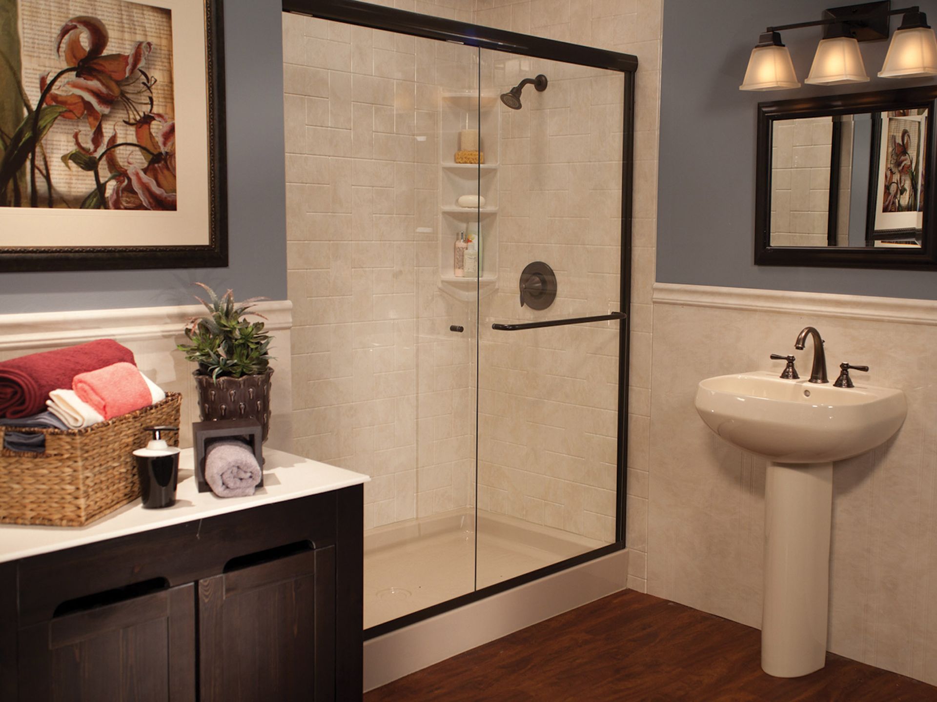 Bathroom with glass shower, pedestal sink, dark cabinet, and decorative shelf on wood floor.