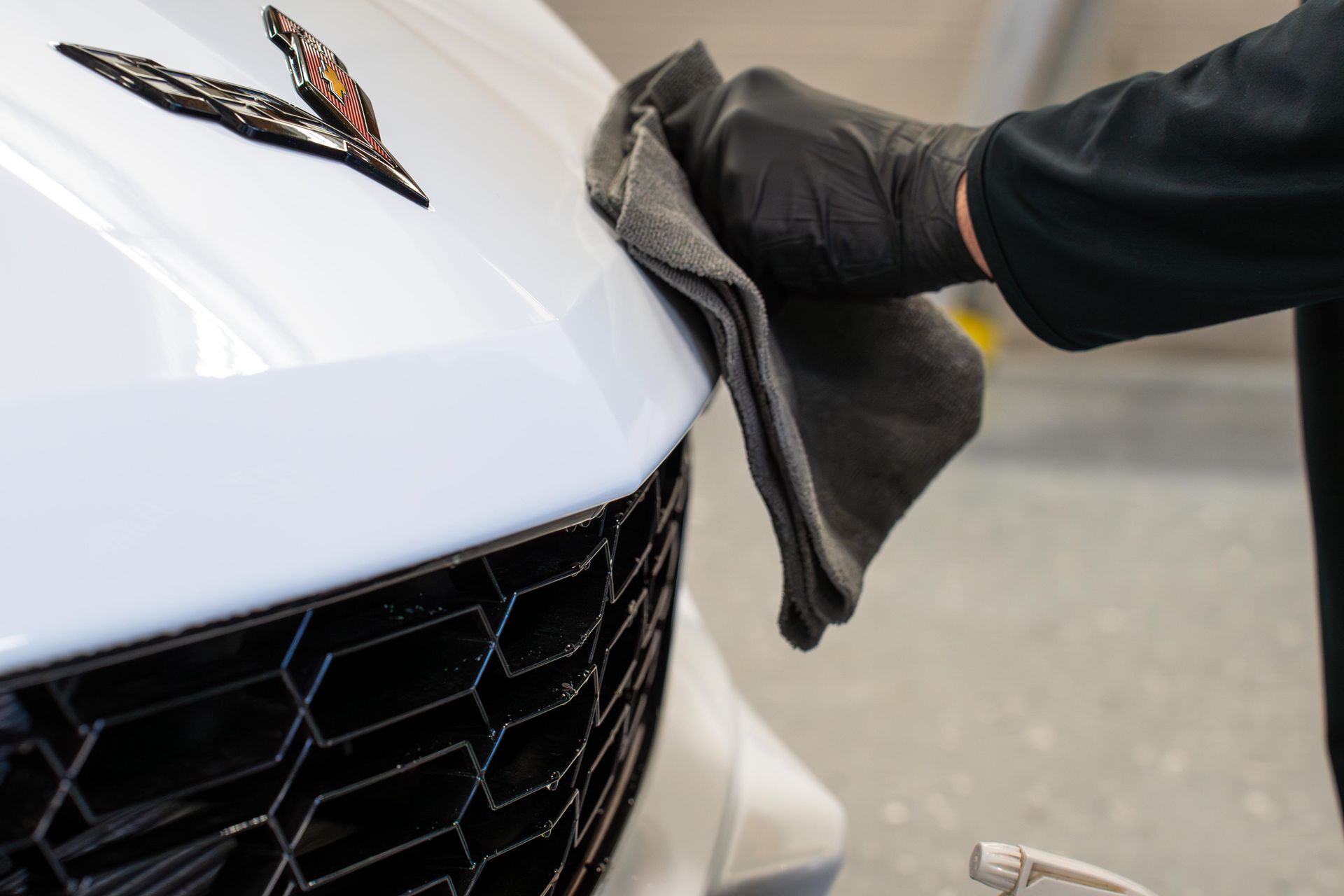 Gloved hand wiping white car hood with gray cloth; close-up shows emblem and black grill.