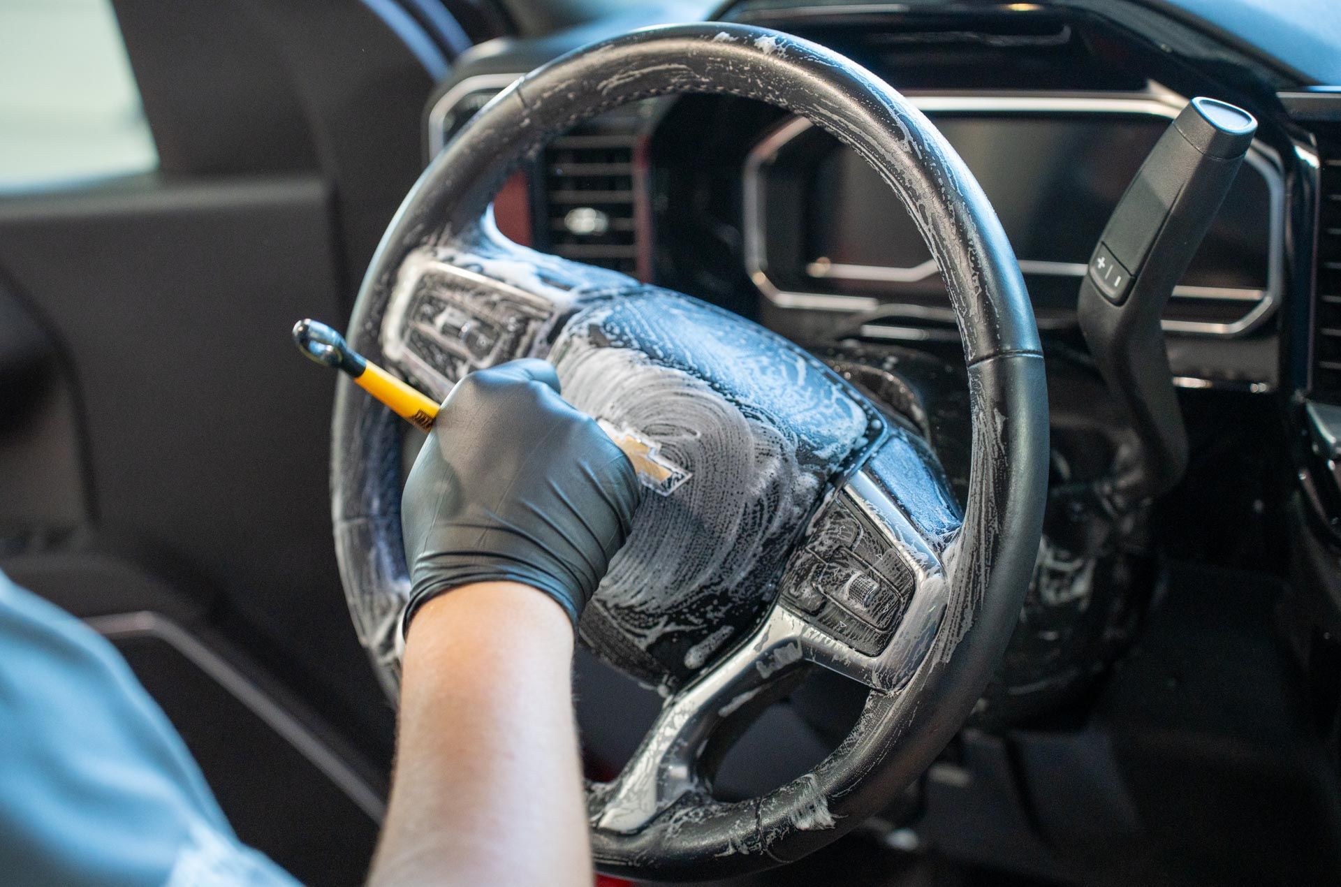 Person wearing black gloves cleaning a car's steering wheel with a brush and soap.