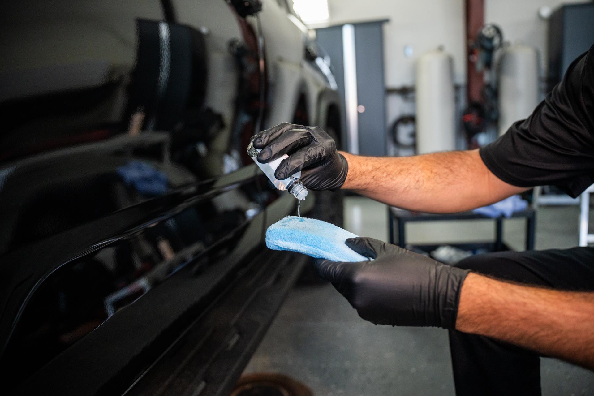 Person applying product to a black car with a sponge in a garage, wearing black gloves.
