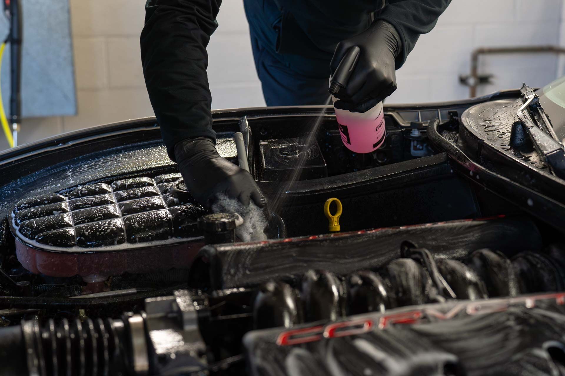 Person cleaning a car engine with a spray bottle and sponge, wearing black gloves.