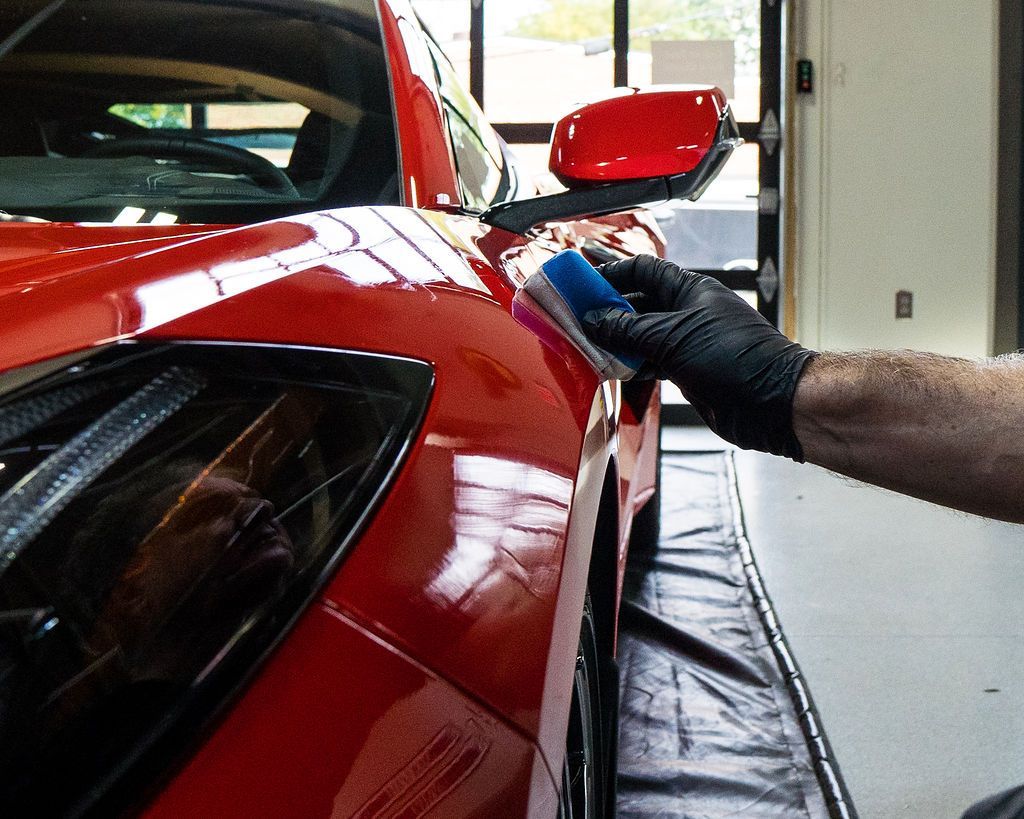 Person applying sealant to a shiny red sports car. They wear a black glove and use a blue applicator. Garage setting.
