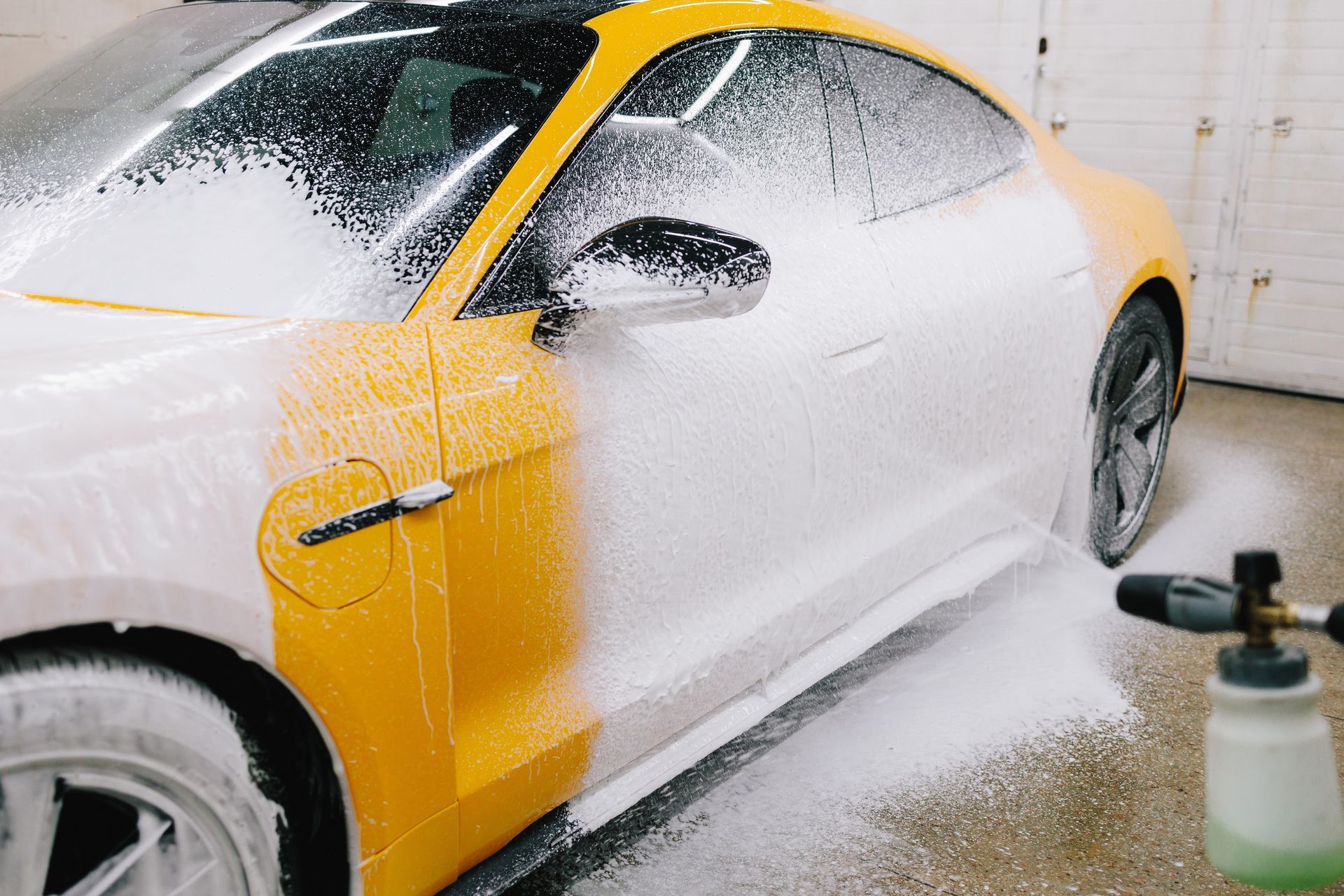 Yellow car covered in white foam, side view, being washed in a garage setting.