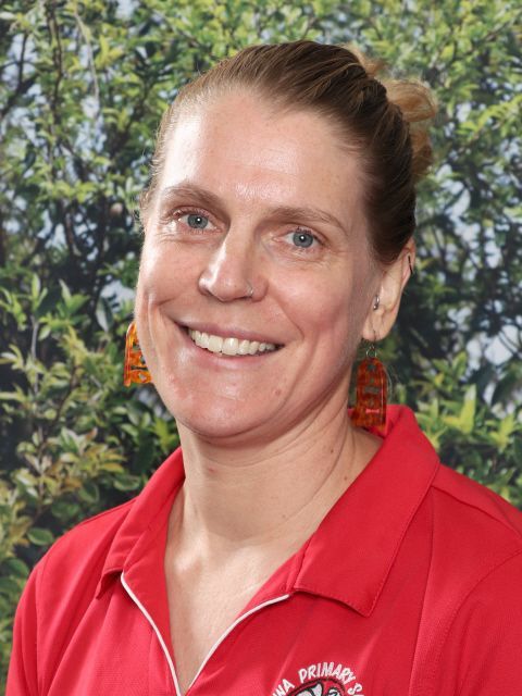 Woman with blonde hair in a red shirt, smiling, set against a leafy background.