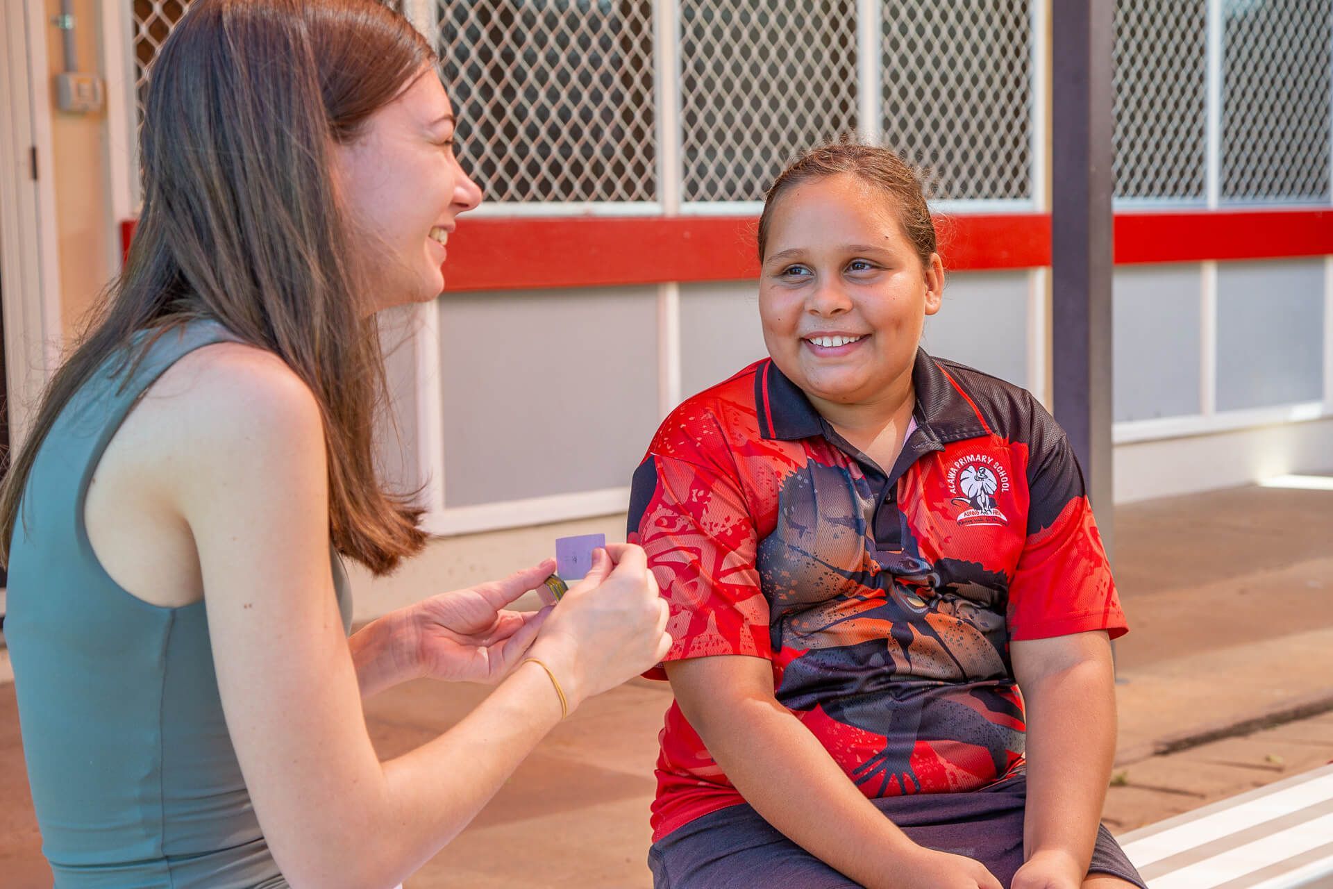 Woman and girl smiling, seated outdoors. Woman holds purple item; girl wears red shirt.