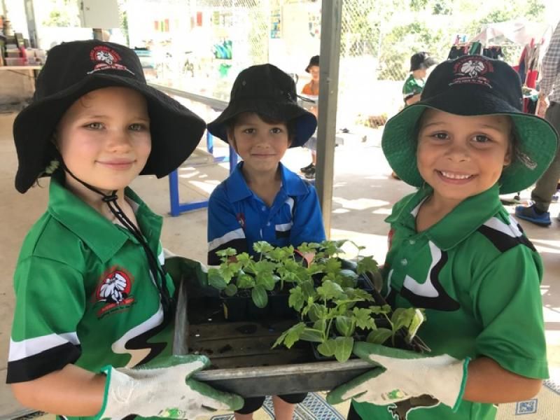 Three children in green shirts and hats hold a tray of seedlings, smiling.