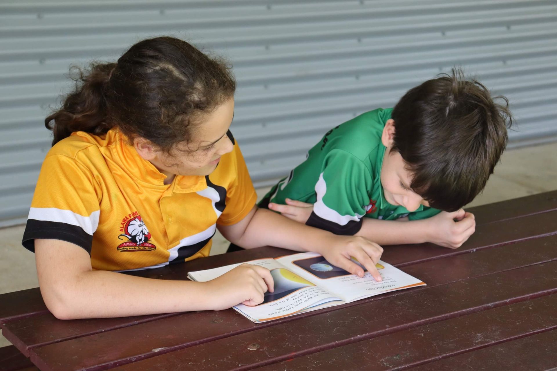 Two children looking at a book on a wooden picnic table; one points at page.