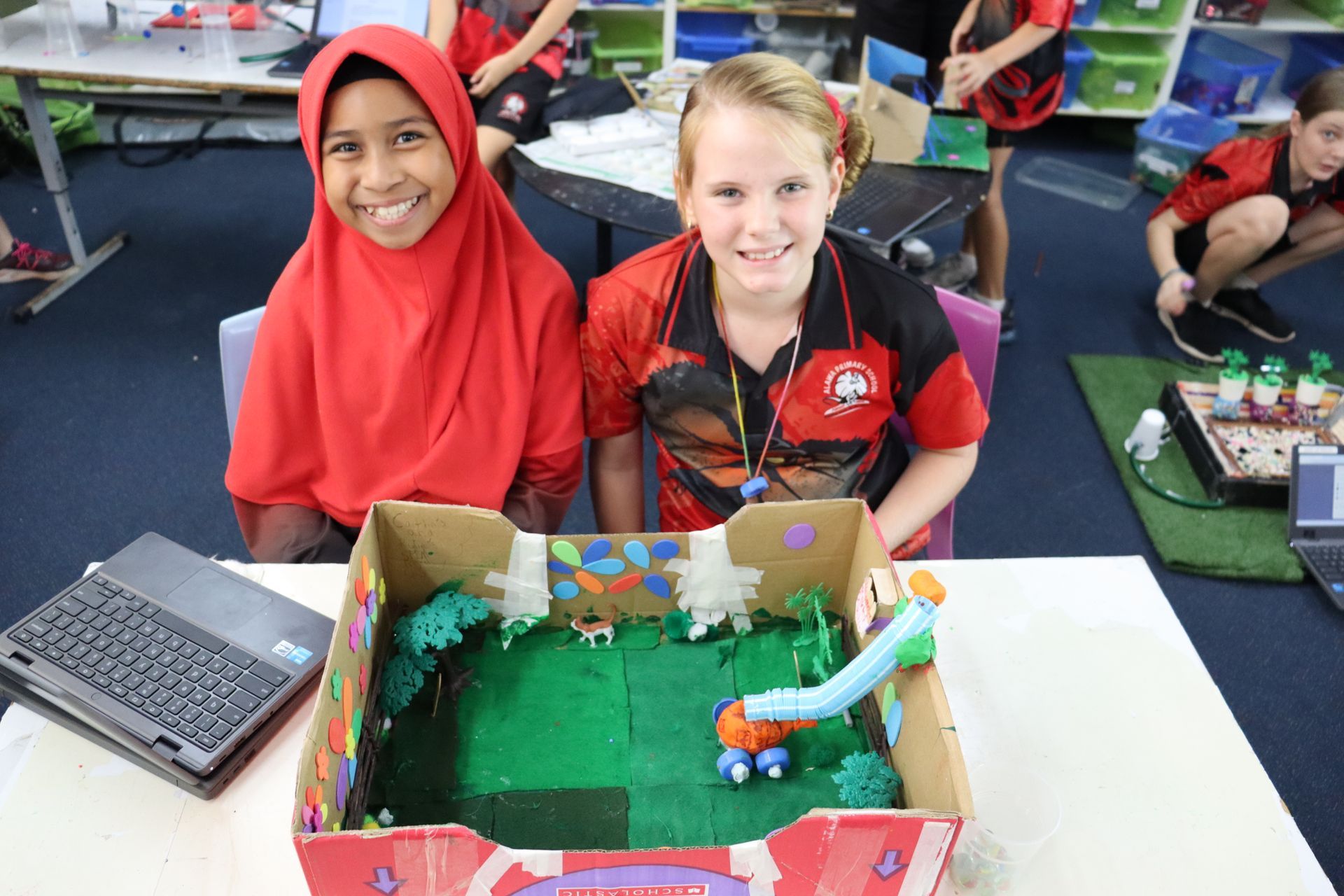 Two girls smiling, showing their project: a model playground in a box.