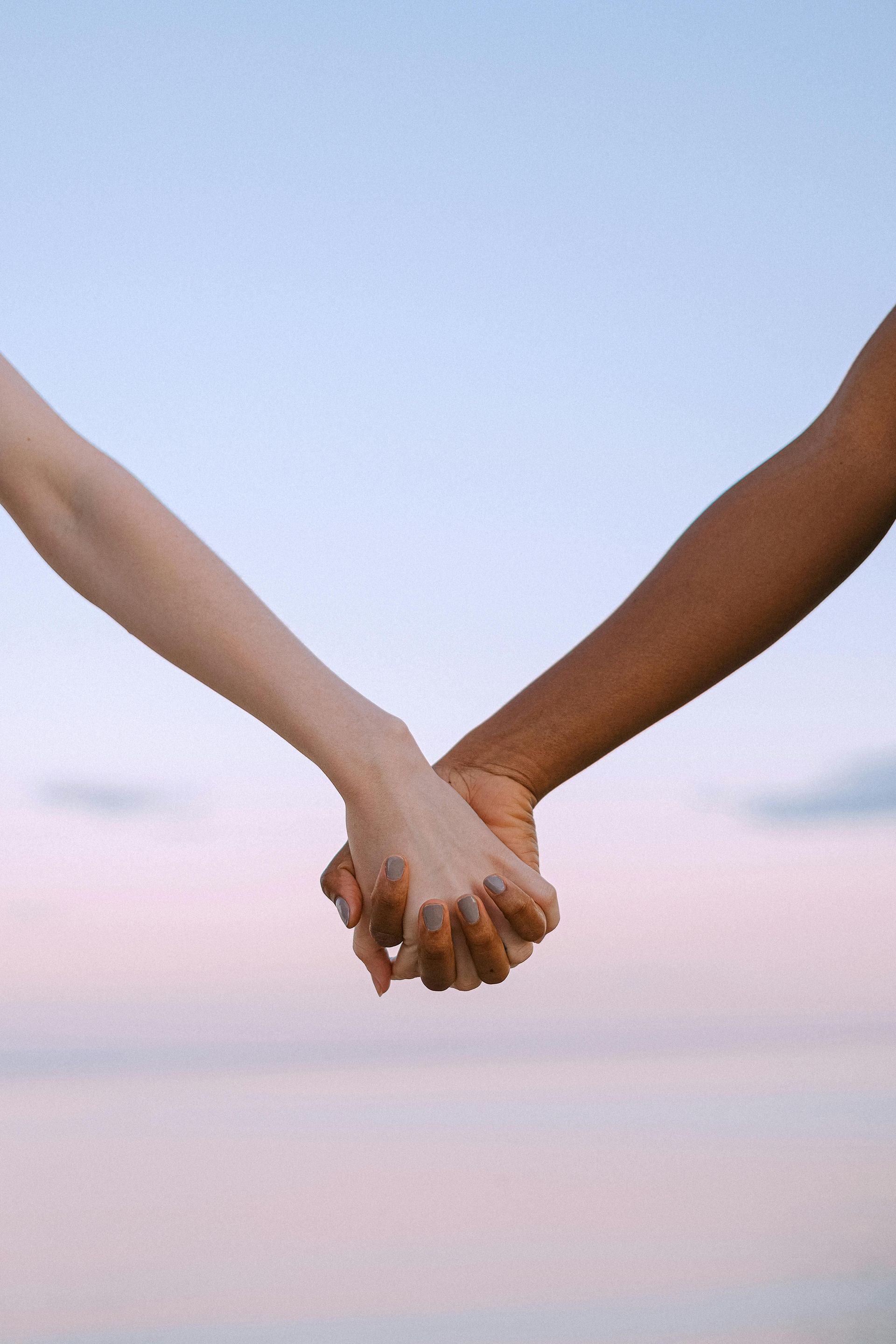 Two hands, one light-skinned and one dark-skinned, clasped together against a pastel sky backdrop.