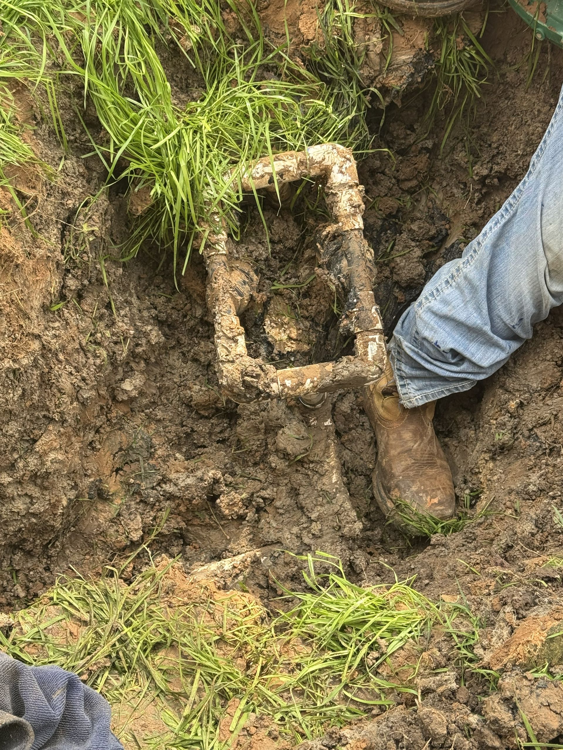 Person digging in muddy ground beside a rusty metal container in a grassy ditch