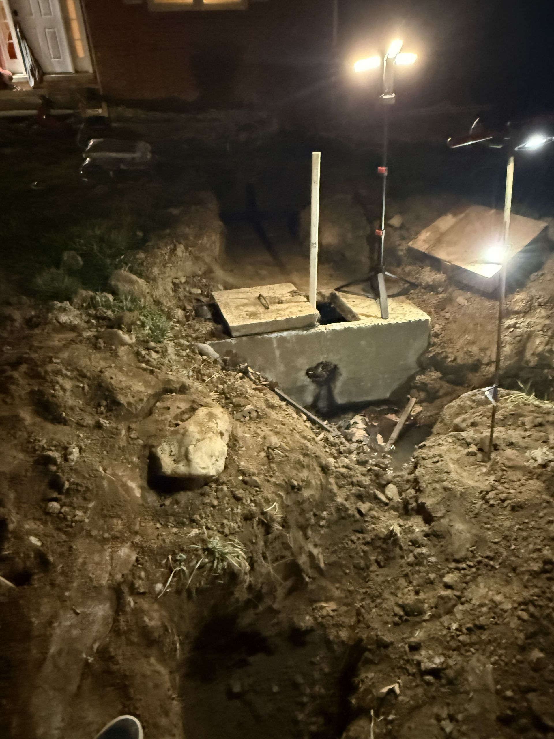 Nighttime construction trench with concrete footing and two work lights illuminating the excavation.