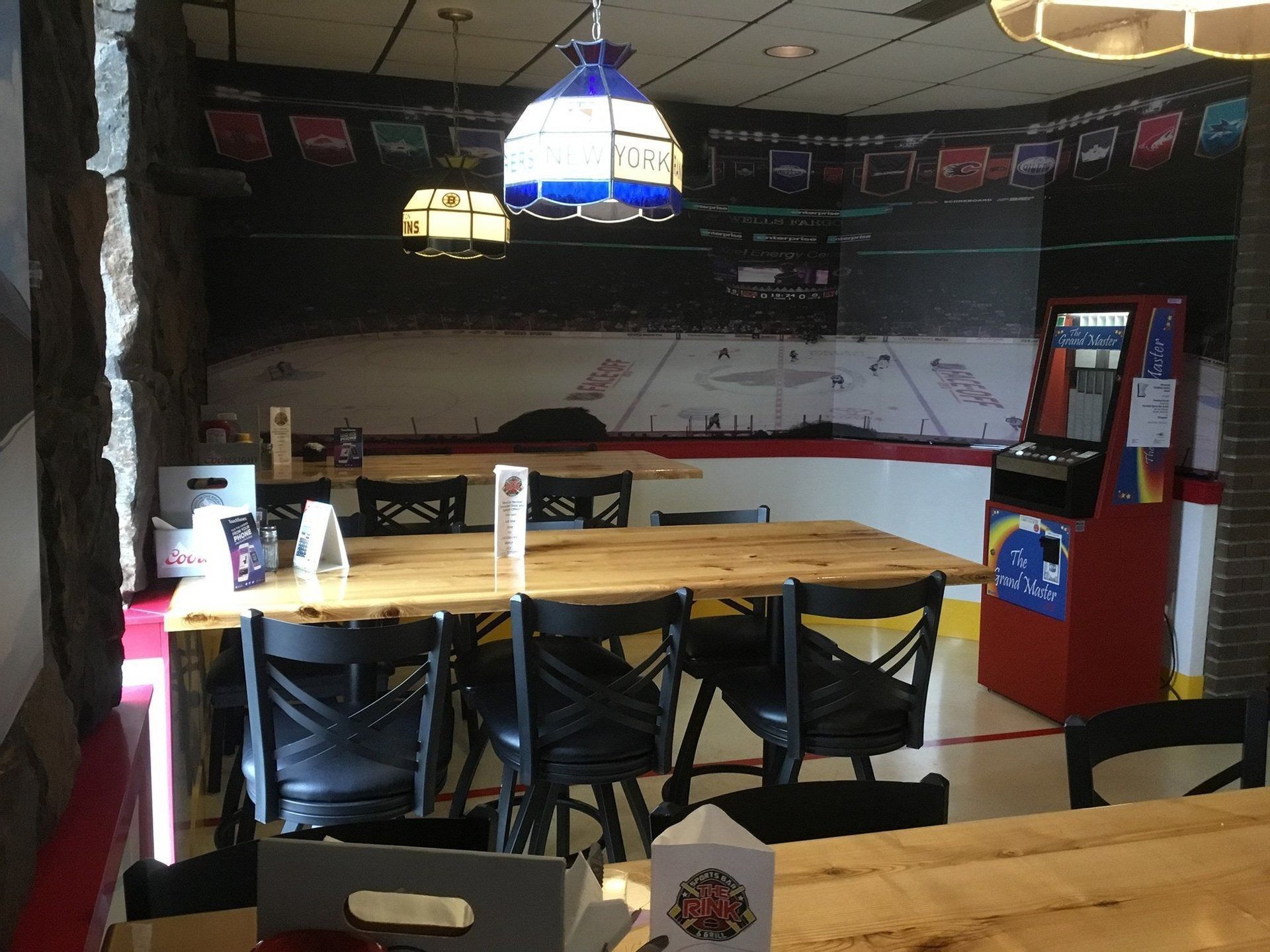Empty sports bar with wooden tables, black chairs, and a blue hanging lamp under a dark mural wall.