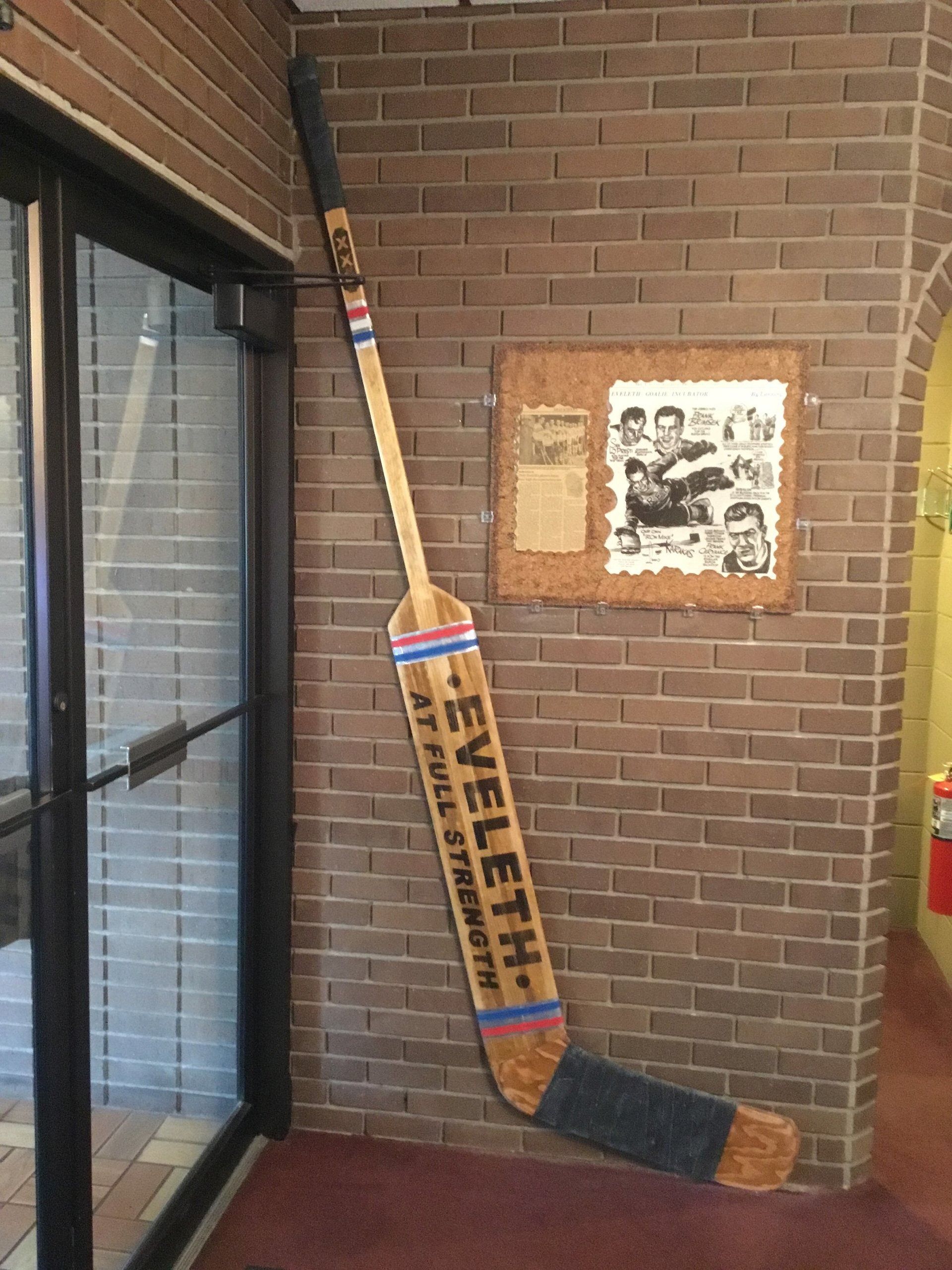 Vintage cricket bat and broom leaning against a brick wall beside a window and framed photo display.