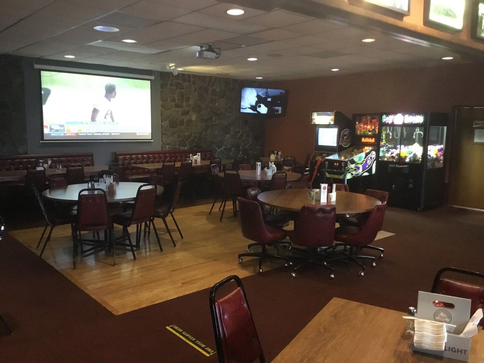 Dim arcade-style bar with large screen, tables, chairs, and vending/game machines in a dark room
