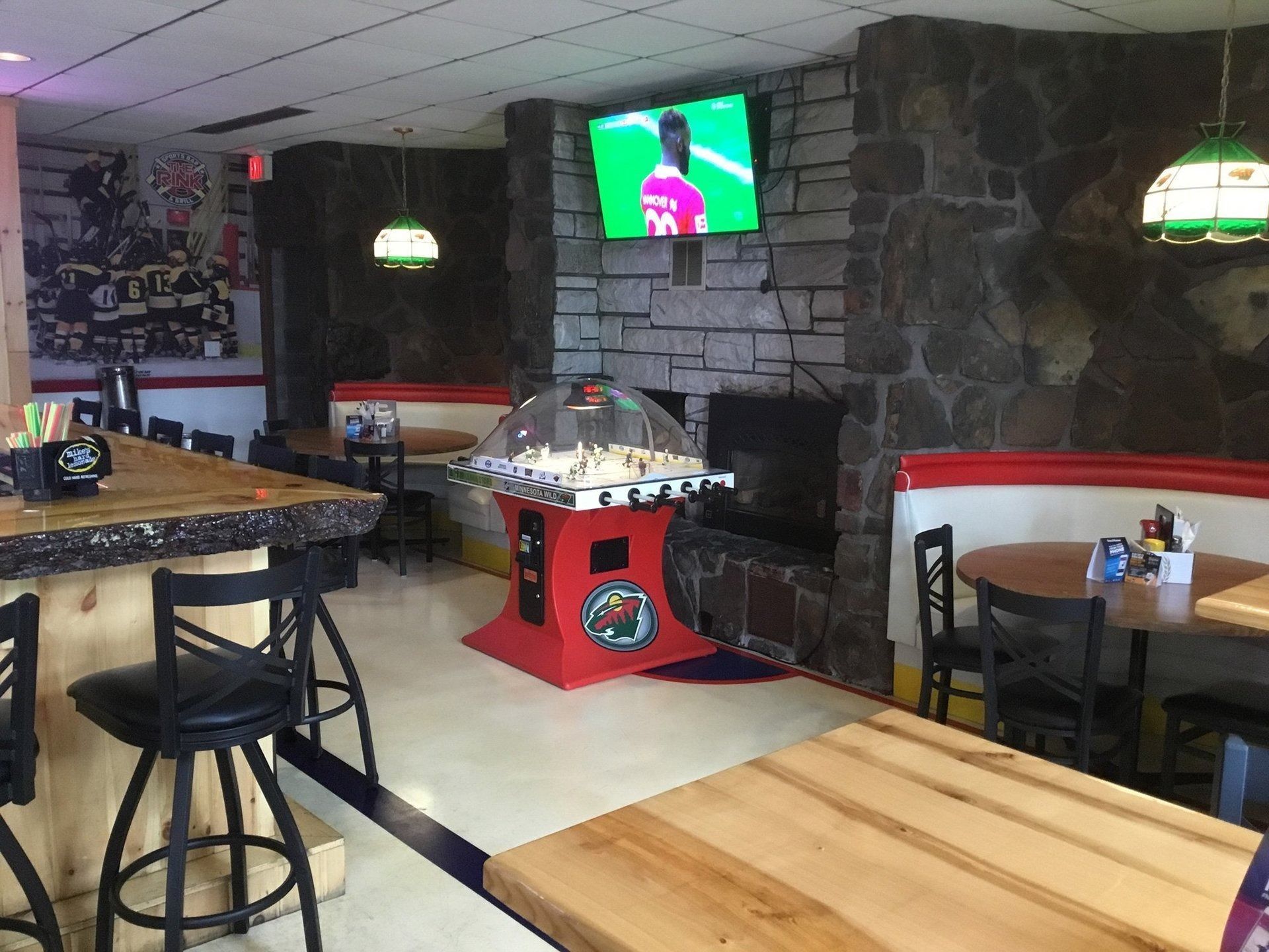 Empty restaurant interior with wooden tables, bar stools, stone fireplace, and a TV on the wall.