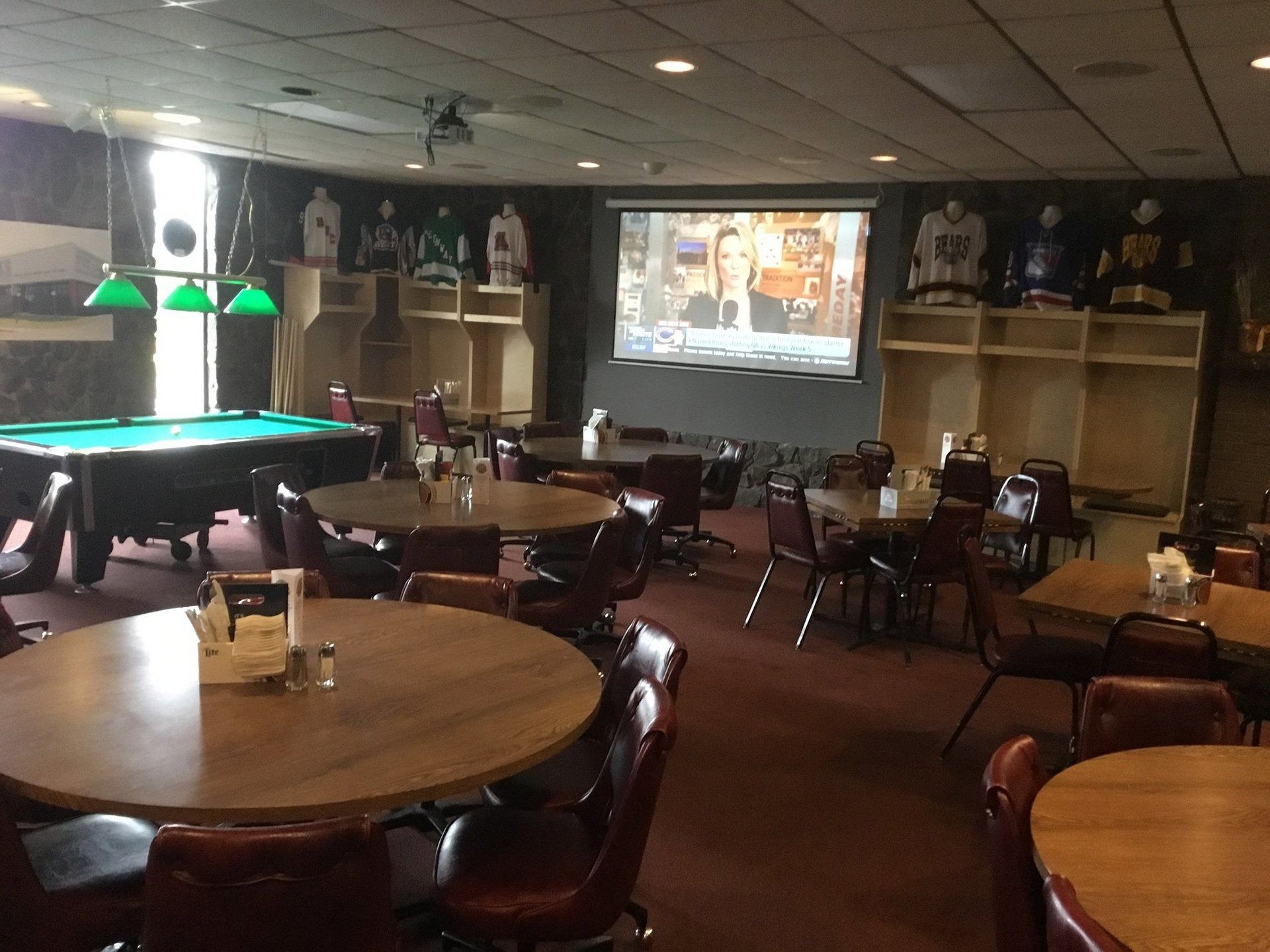 Dim lounge with round tables, pool table, bar, and a projection screen showing a game.