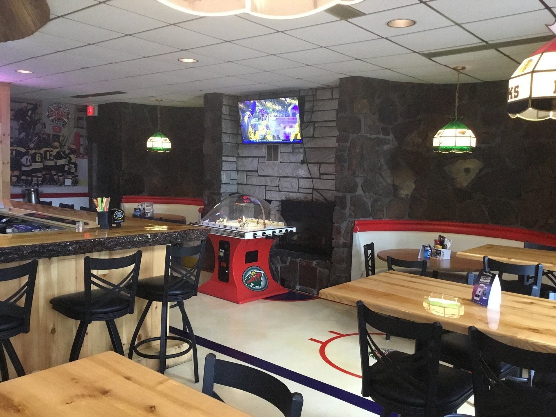 Cozy restaurant interior with wooden tables, black stools, stone wall, and a TV above a red arcade game.