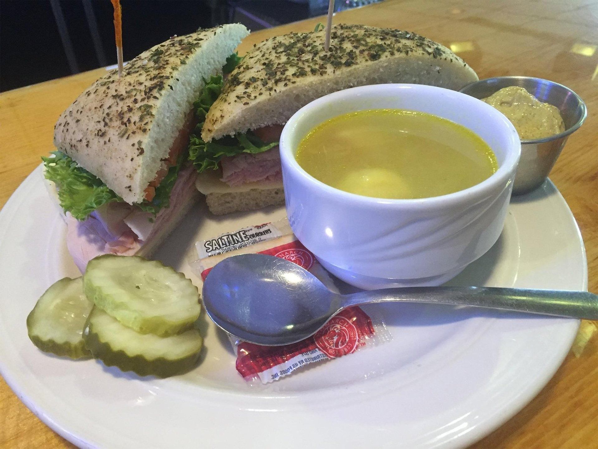 Sandwich on a plate with soup, pickles, and a spoon on a table.