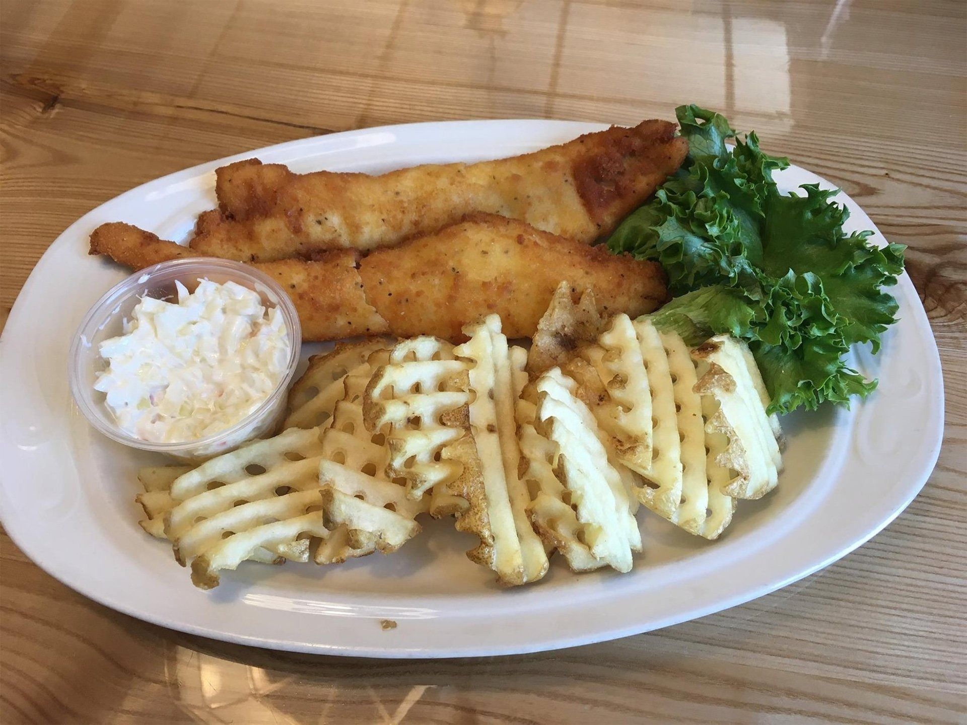 Plate of fried fish with waffle fries, coleslaw, and parsley garnish on a wooden table