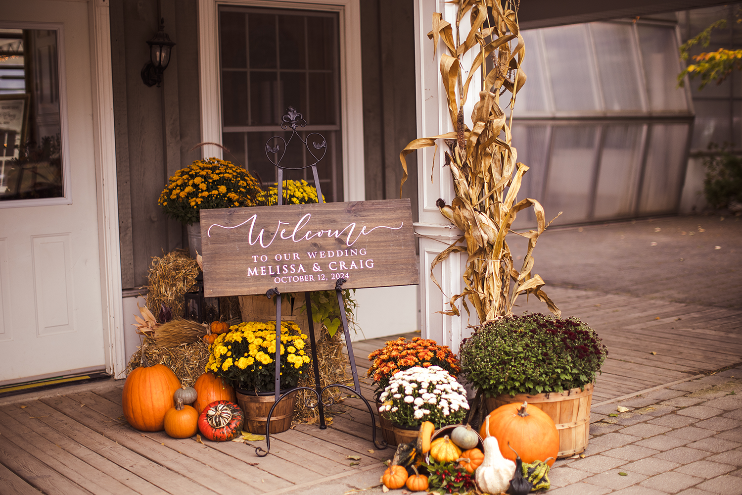 A welcome sign is sitting on a porch surrounded by pumpkins and flowers.