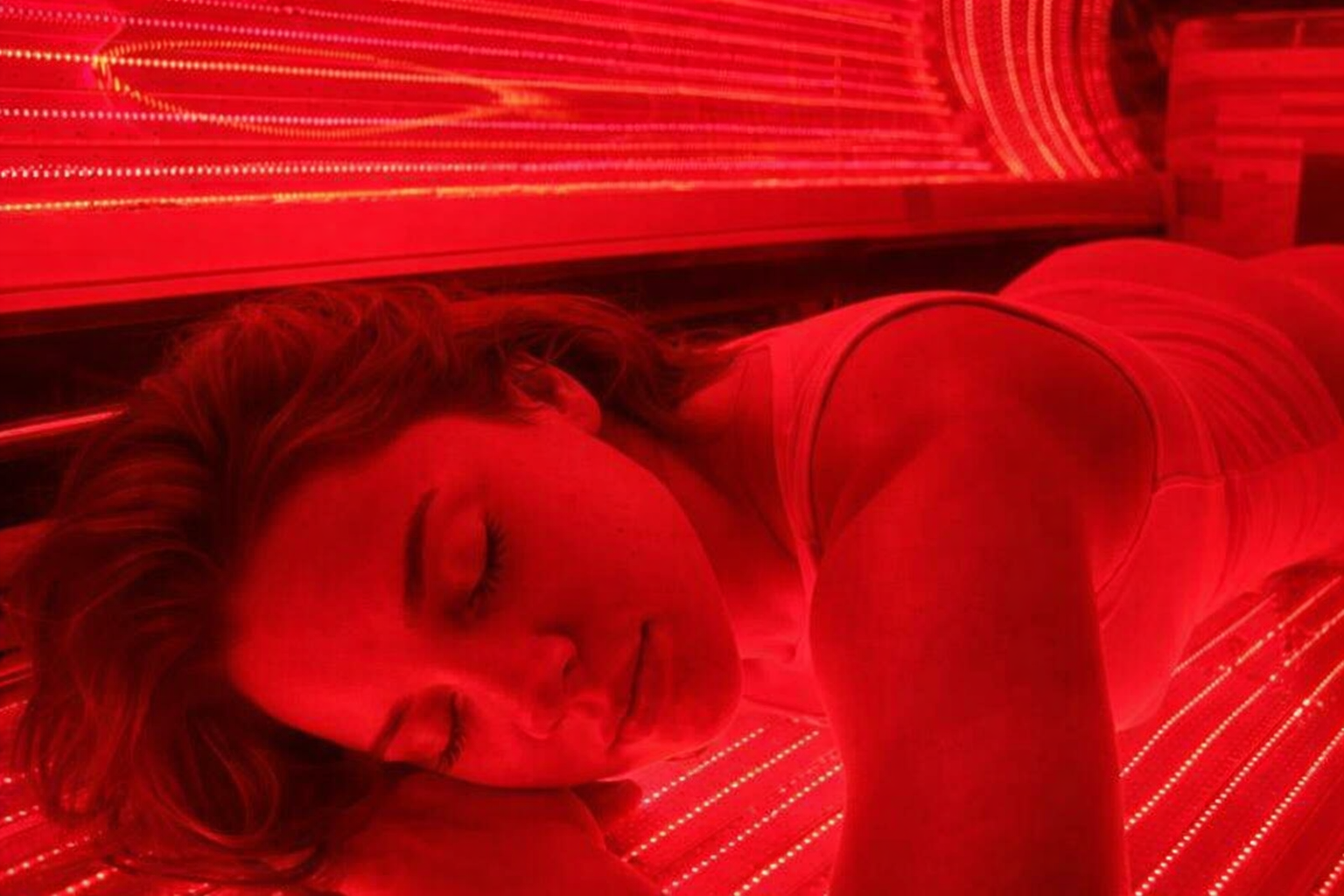 Woman lying inside a red-lit tanning bed with eyes closed
